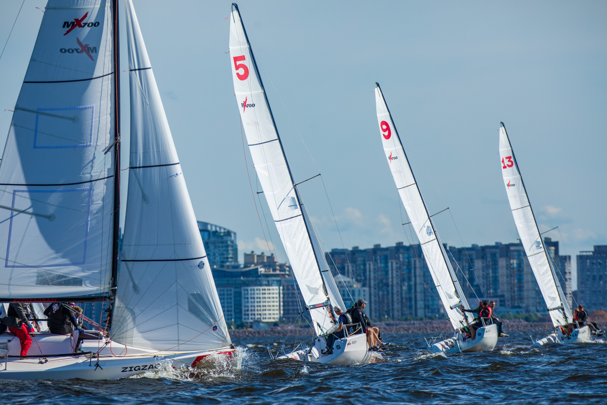 Парусные яхты идут симметрично на фоне жилых домов Петербурга.