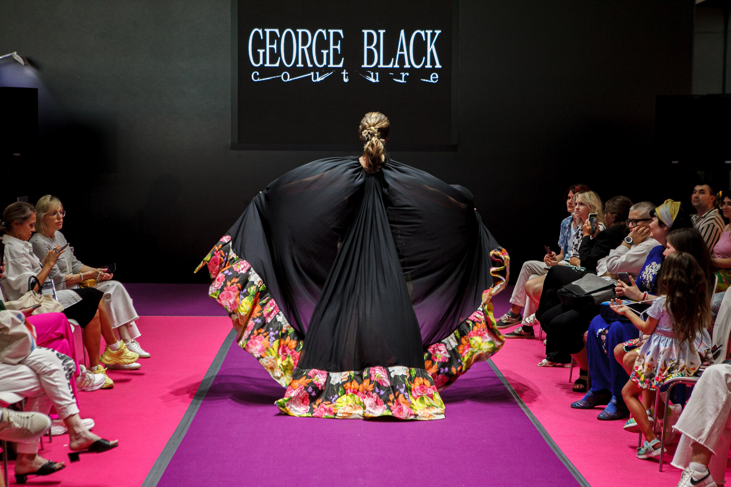  "Модели в ярких нарядах проходят по подиуму во время показа мод, освещенные яркими софитами и окруженные восторженными зрителями."