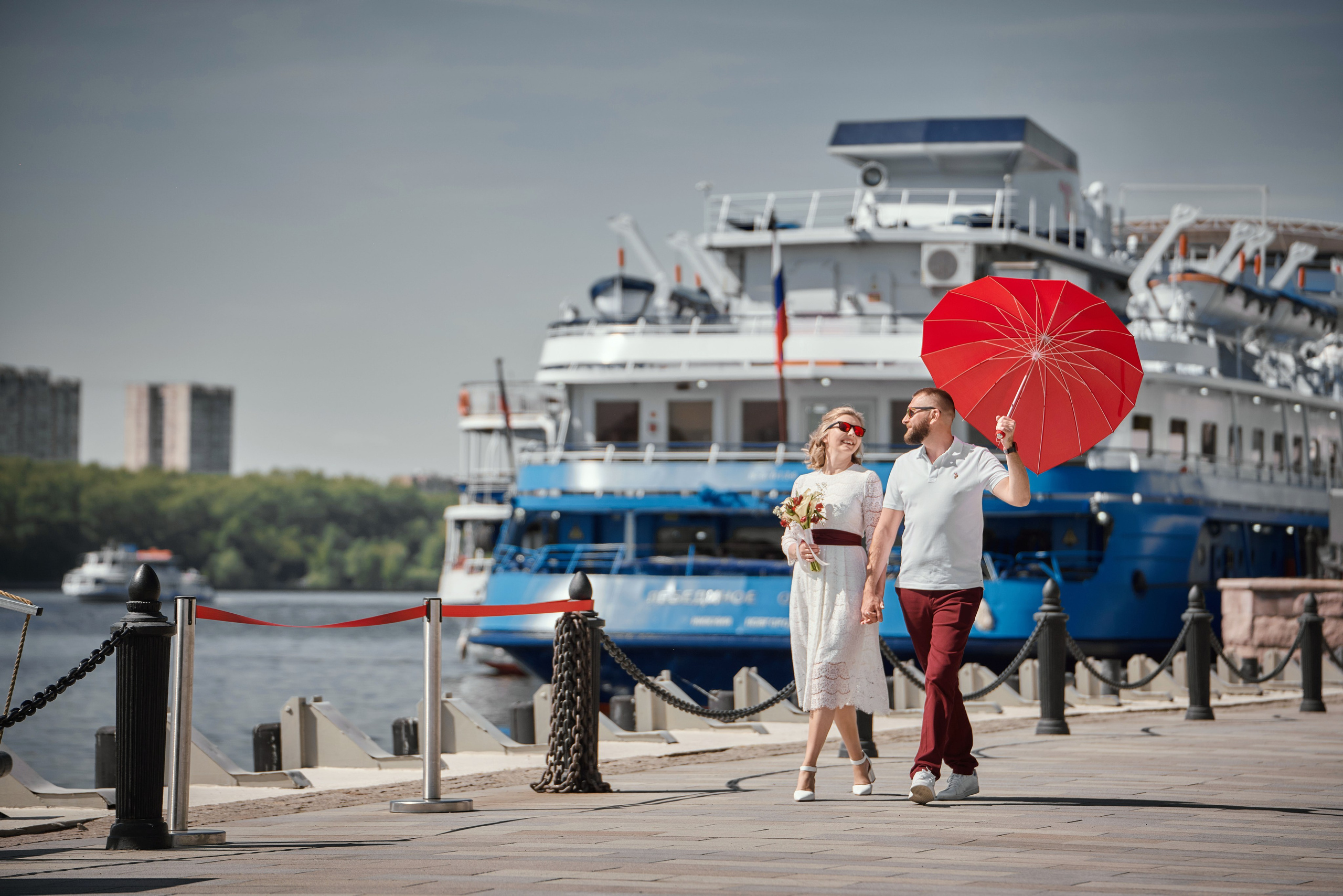 жених и невеста на свадебной фотосессии идут по набережной Москвы реки у речного вокзала с красным зонтом
