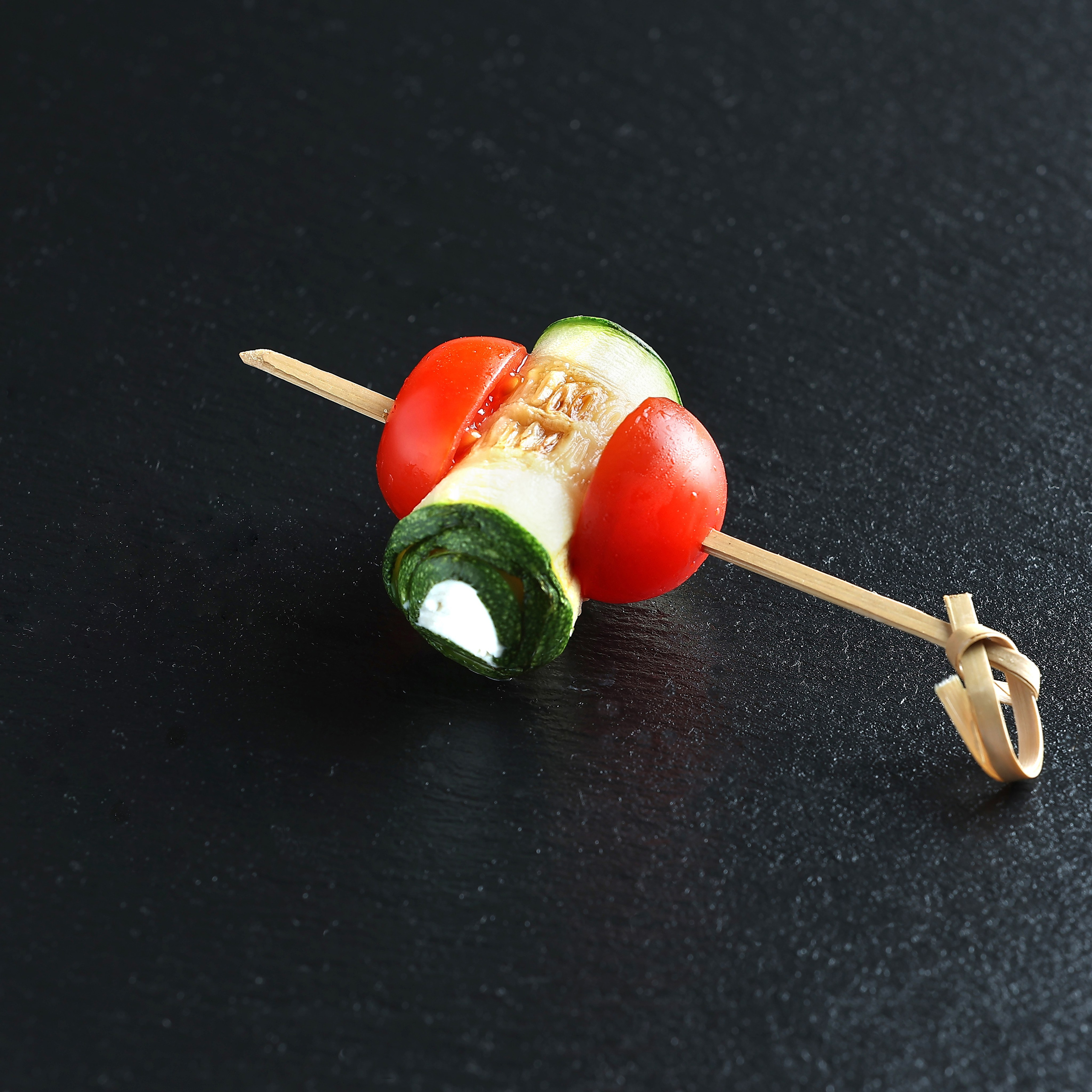 Закуска на шпажке: завиток запечённого цукини с крем-сыром между половинками помидора черри, на тёмном фоне.