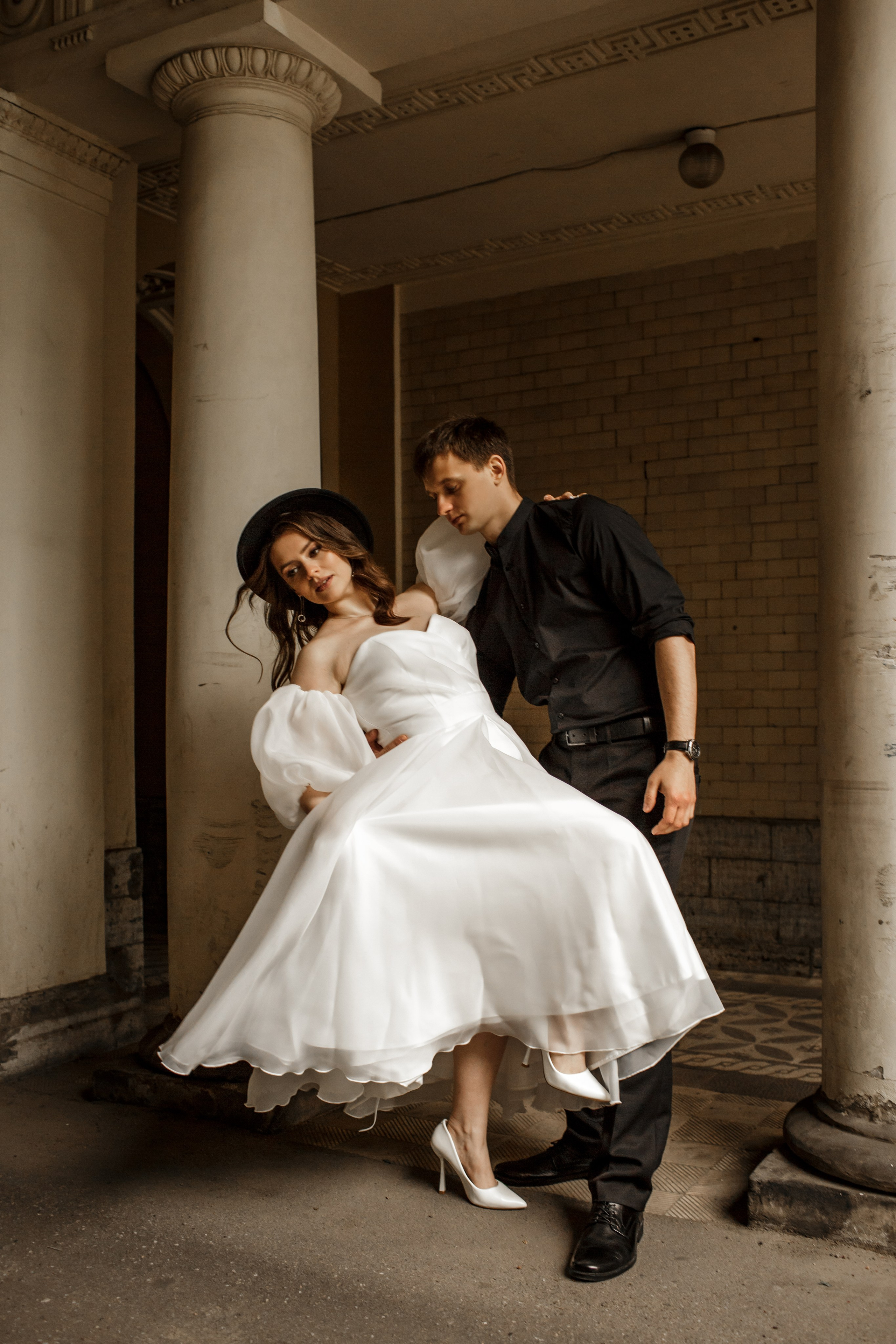 Молодожены в Санкт-Петербурге во дворе с колоннами; жених держит невесту за талию; невеста поднимает подол белого платья.
