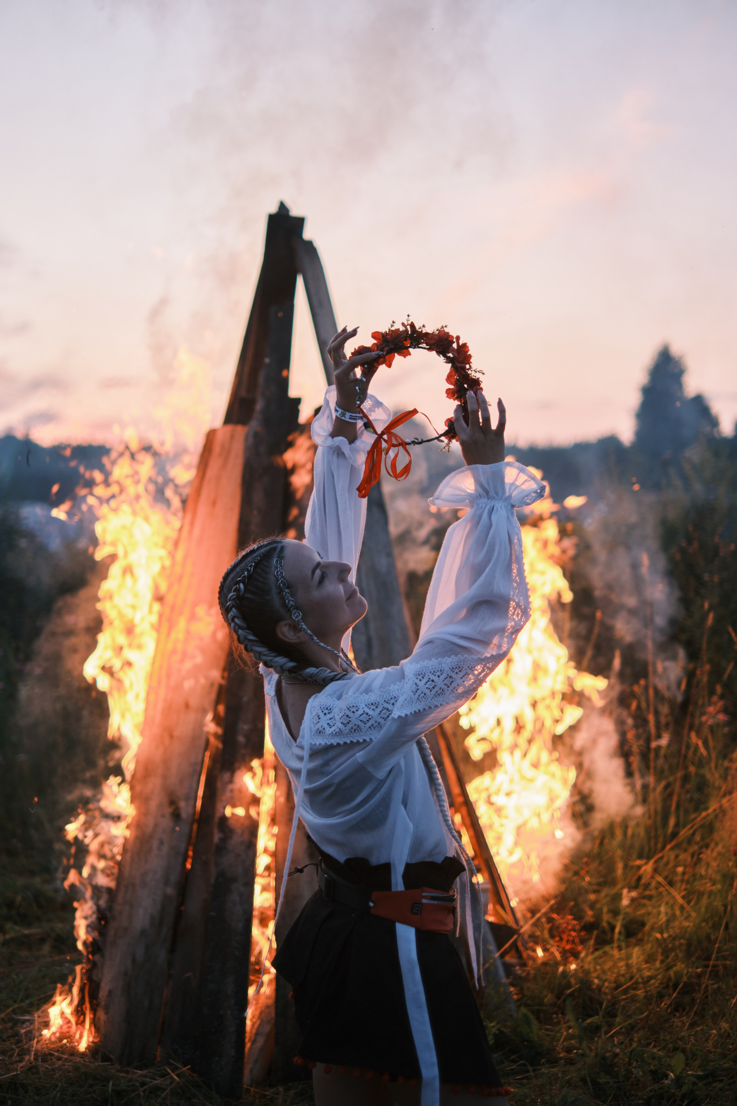 Девушка стоит в русском костюме вытягивает руки вверх держа венок над большим костром