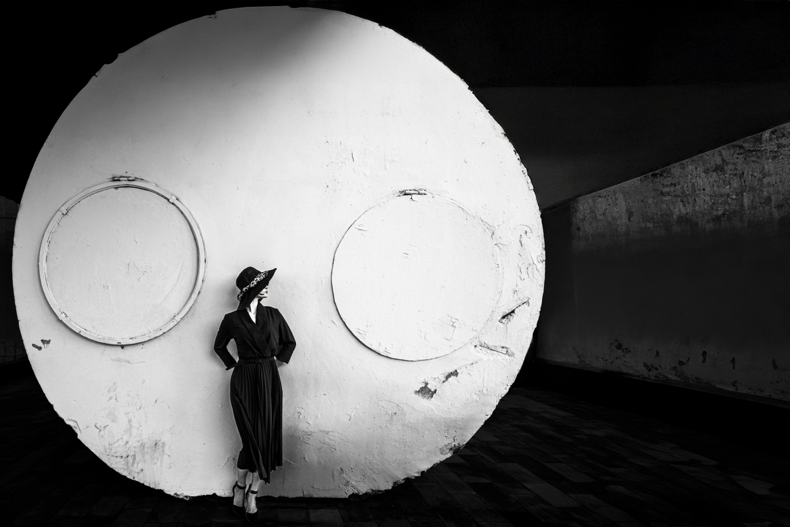 Женщина в чёрном платье и шляпе позирует у гигантского круглого объекта на фоне графичной архитектуры. Чёрно-белая арт-фотография.