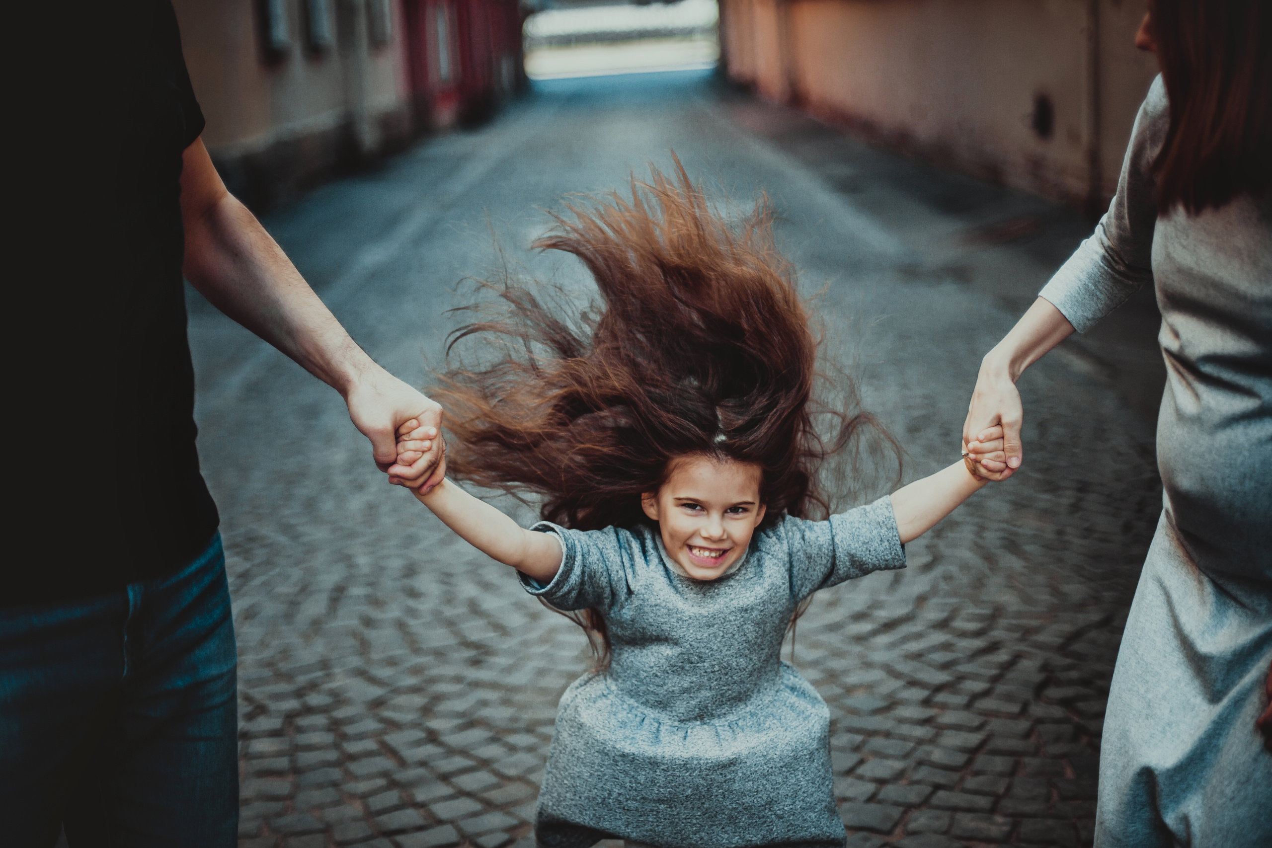 родители держат дочку с развивающимися волосами за руки на фоне брусчатки улицы Репина