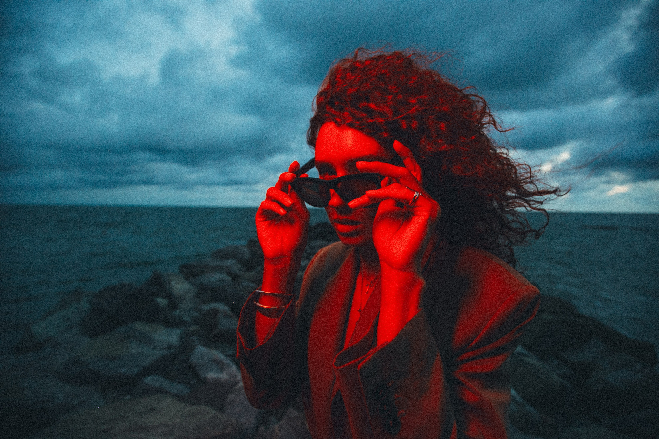 Драматичный портрет девушки с кудрявыми волосами на фоне вечернего моря и грозового неба. Лицо и фигура залиты интенсивным красным светом. Фотограф: Галина Скорик (Сочи).