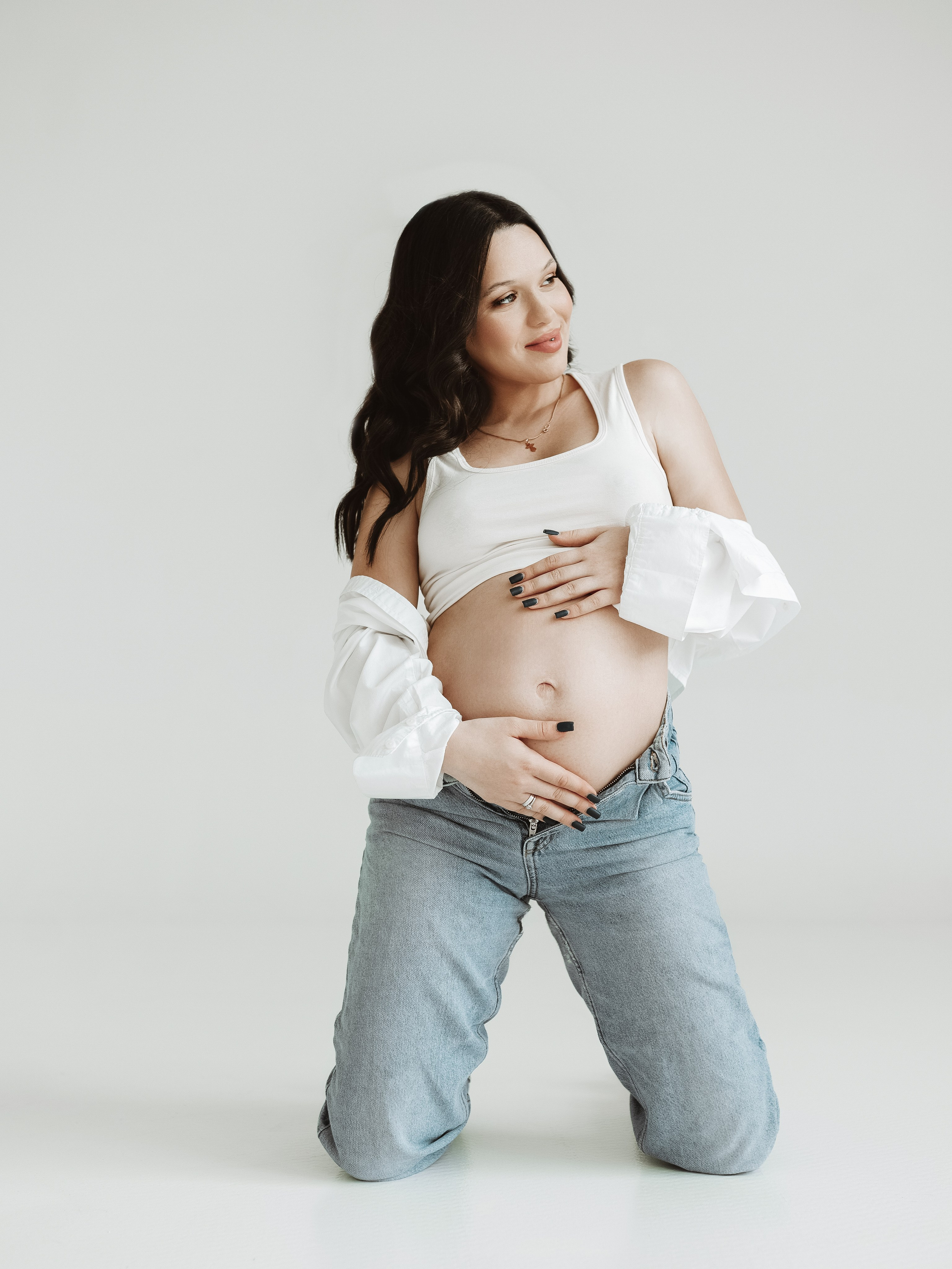 Лёгкий минимализм: будущая мама в белой майке и джинсах позирует на коленях в светлой студии Новосибирска; естественная атмосфера и подчёркнутый живот — фирменный стиль фотографа Яны Ивановой.