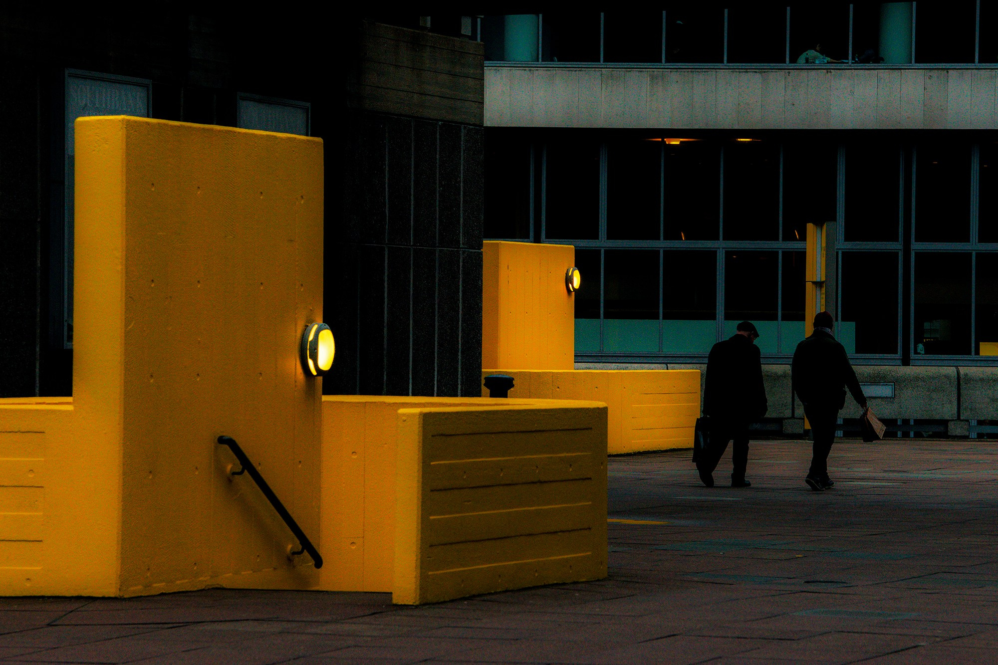 Ярко-желтые бруталистские бетонные конструкции и ступени Хейворд-Гэллери в Лондоне, освещенные фонарями на фоне темного здания и двух прохожих, динамичные тени