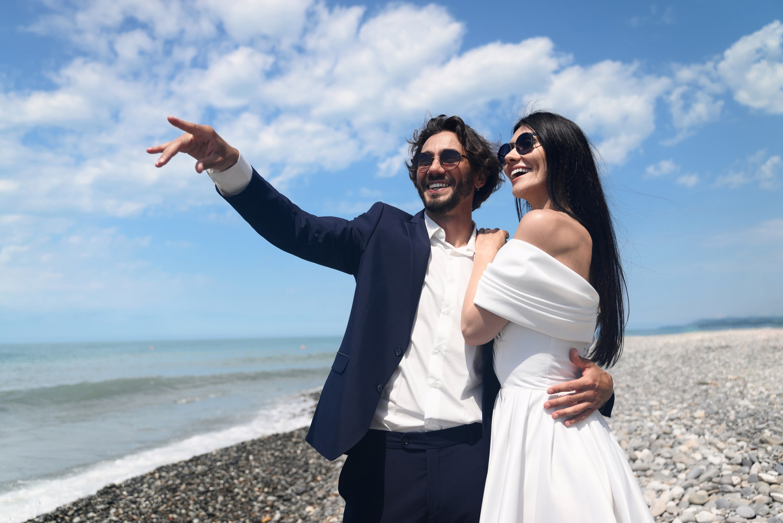 Нежная фотография где двое влюблённых сердца которые только только обменялись кольцами стоят на берегу моря в городе Сочи и смотрят куда-то вдаль жених что-то показывает невесте как будто увидел в море что-то