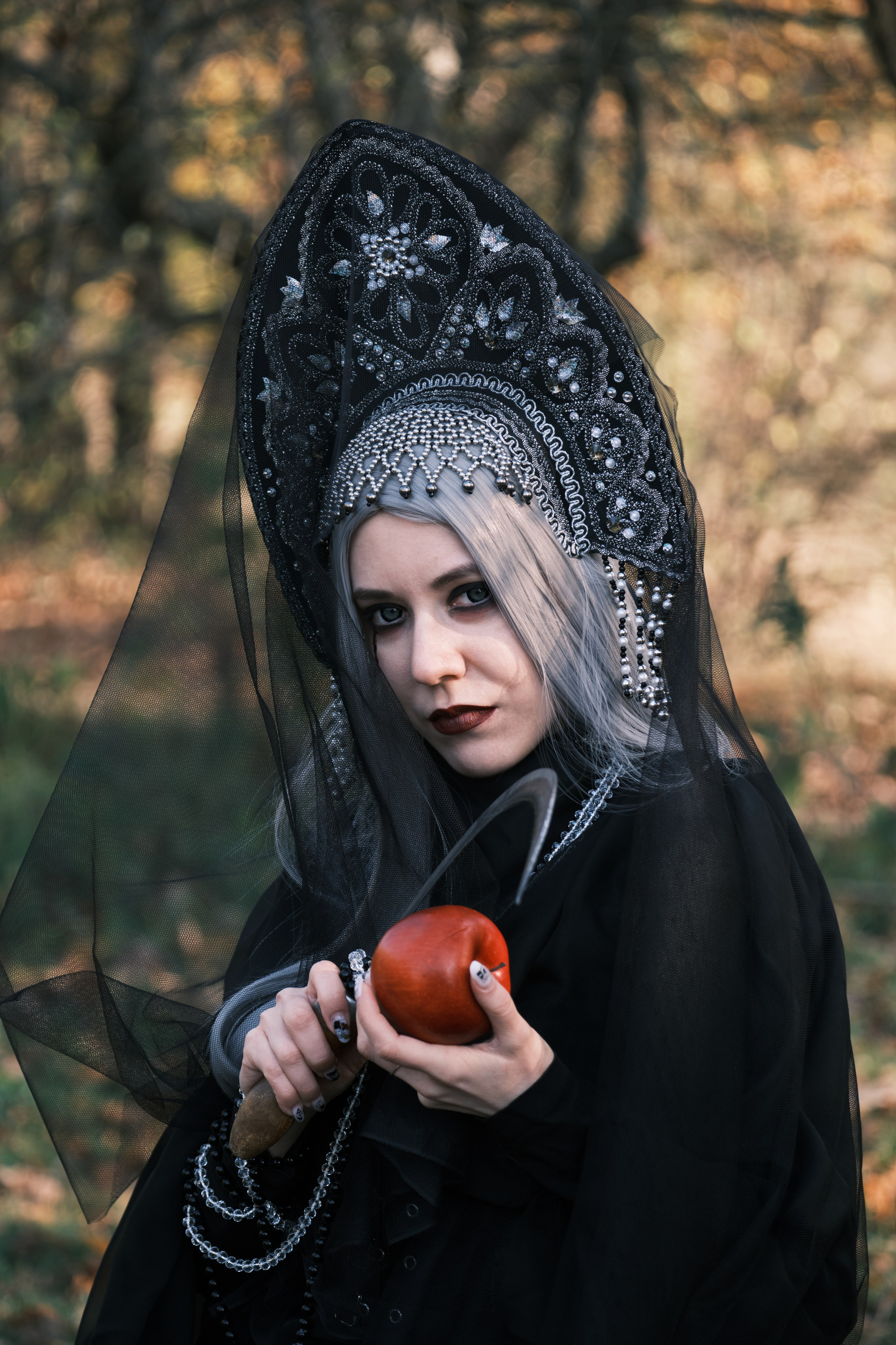 Девушка в готическом черном наряде с кокошником режет яблоко серпом