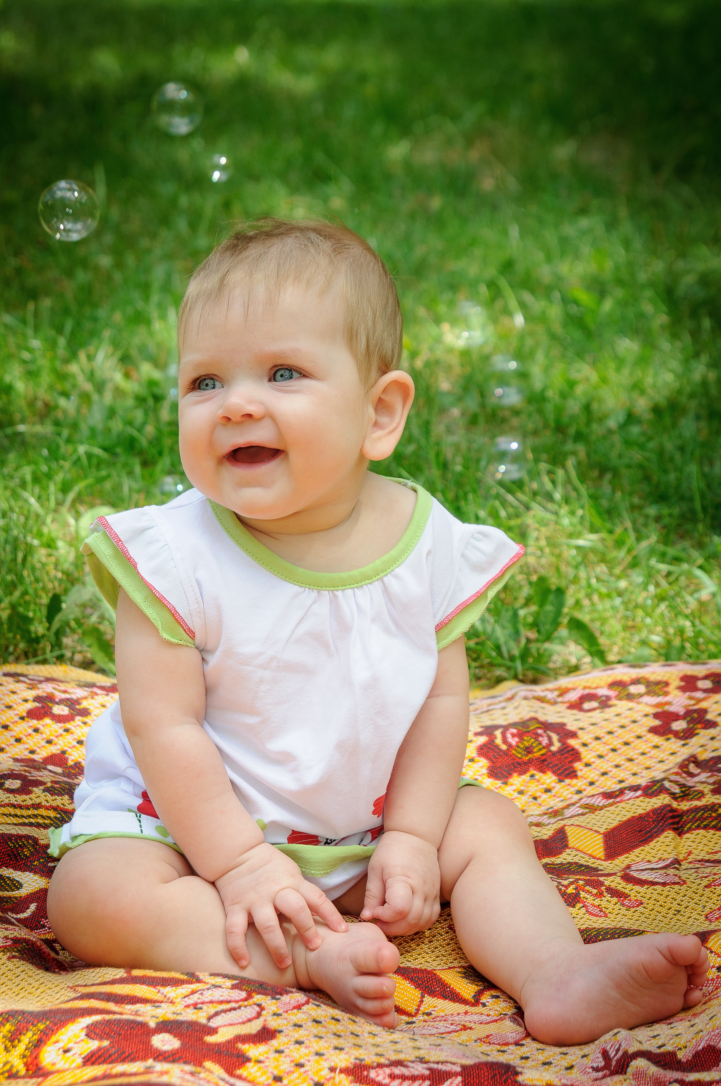 Ребенок улыбается сидя на коврике на траве с мыльными пузырями вокруг