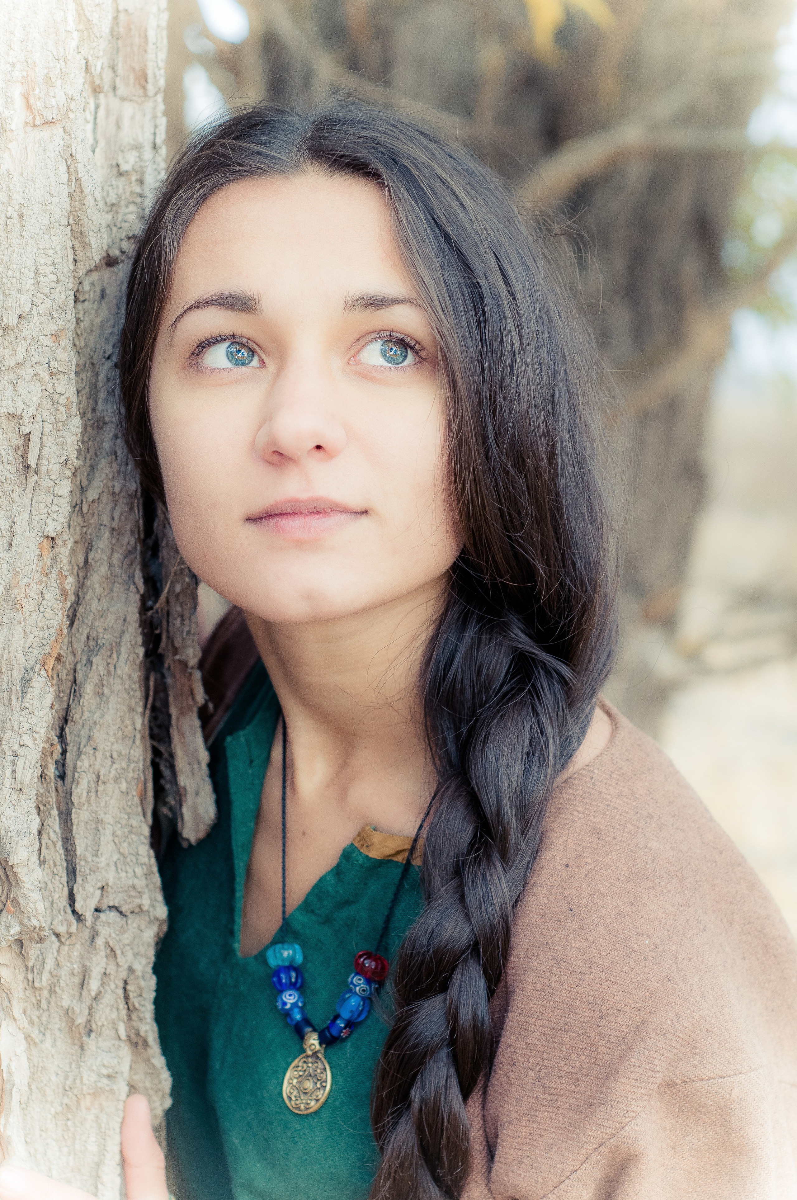 Девушка с косой и украшением на шее стоит около дерева