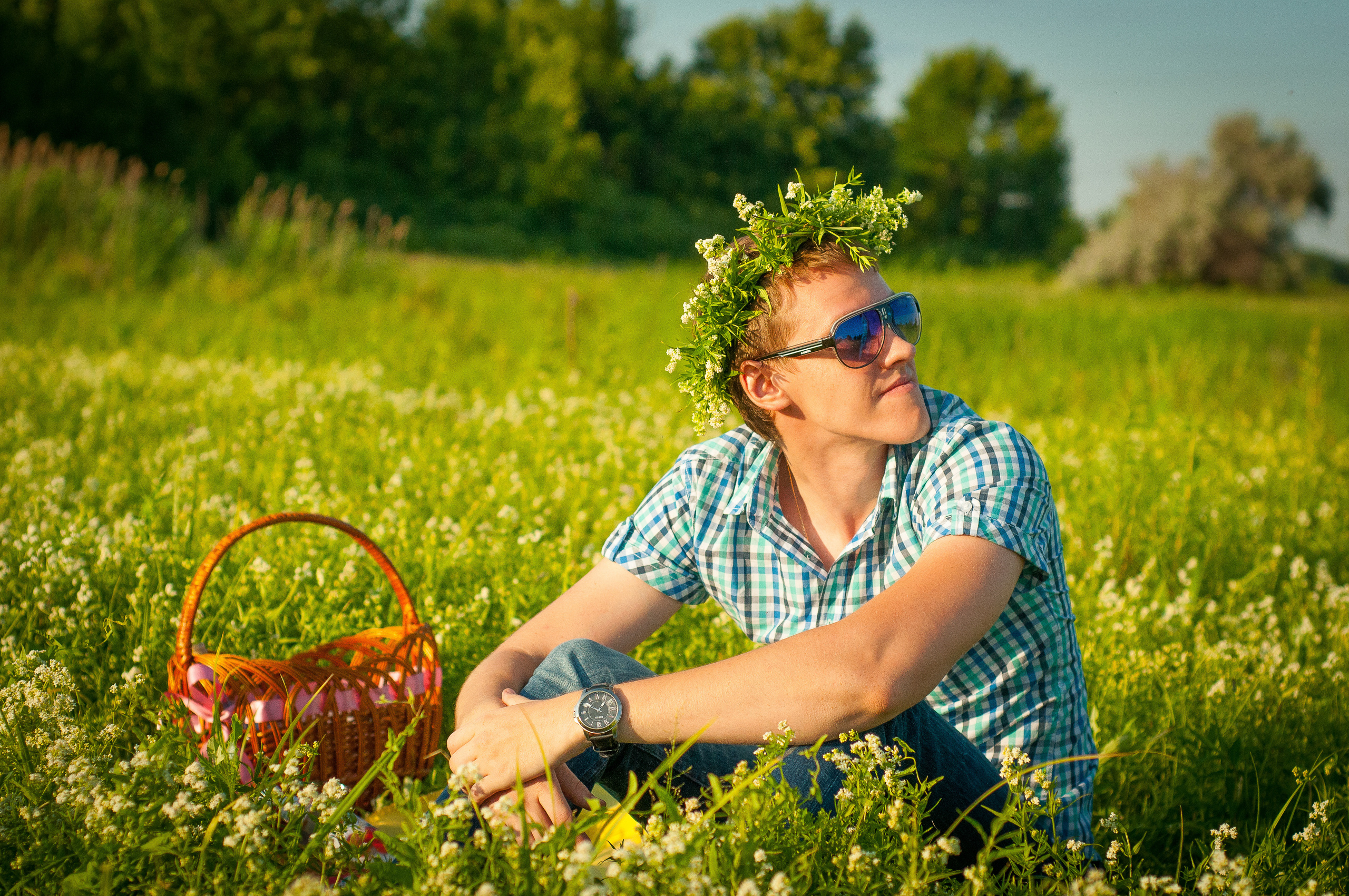 Парень в очках и венком на голове сидит в летнем поле с корзинкой