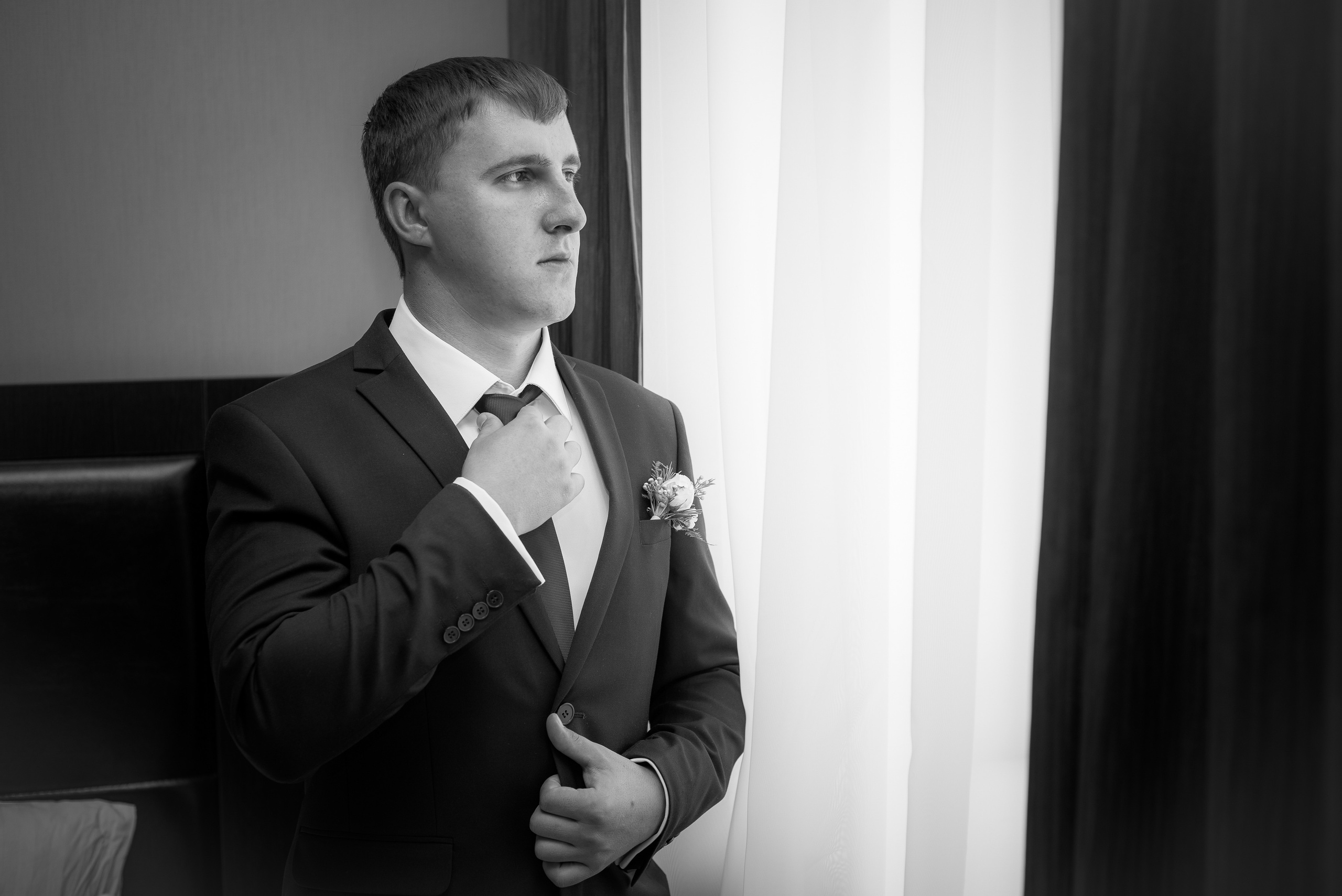 Жених стоит в чёрном костюме около окна и поправляет галстук