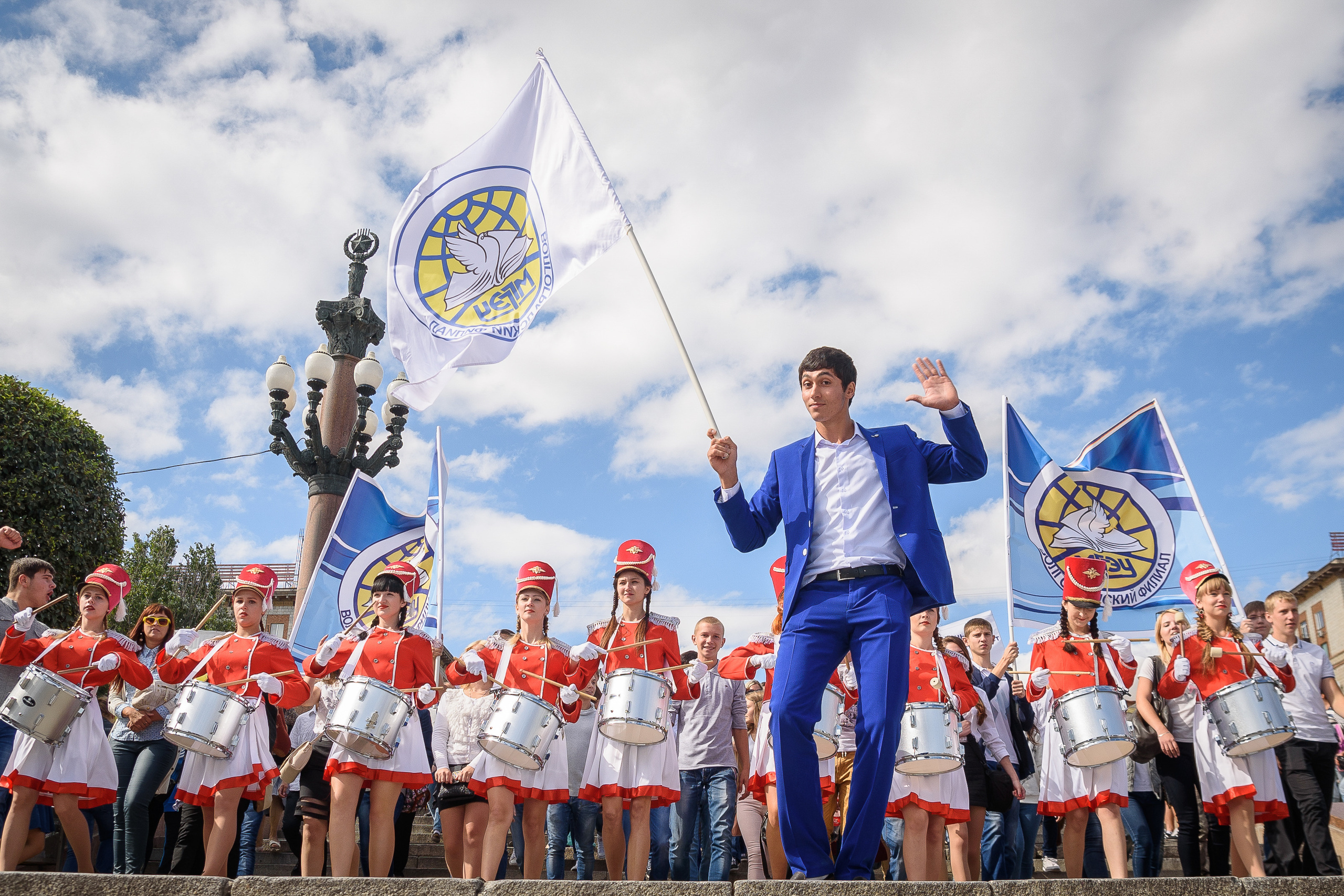 Парень с синем костюме с флагом и барабанщицами на фоне