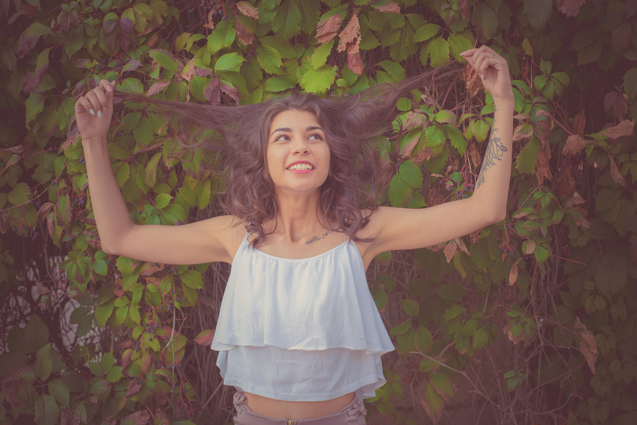 Девушка в белом топе держит себя за волосы на фоне виноградных зарослей