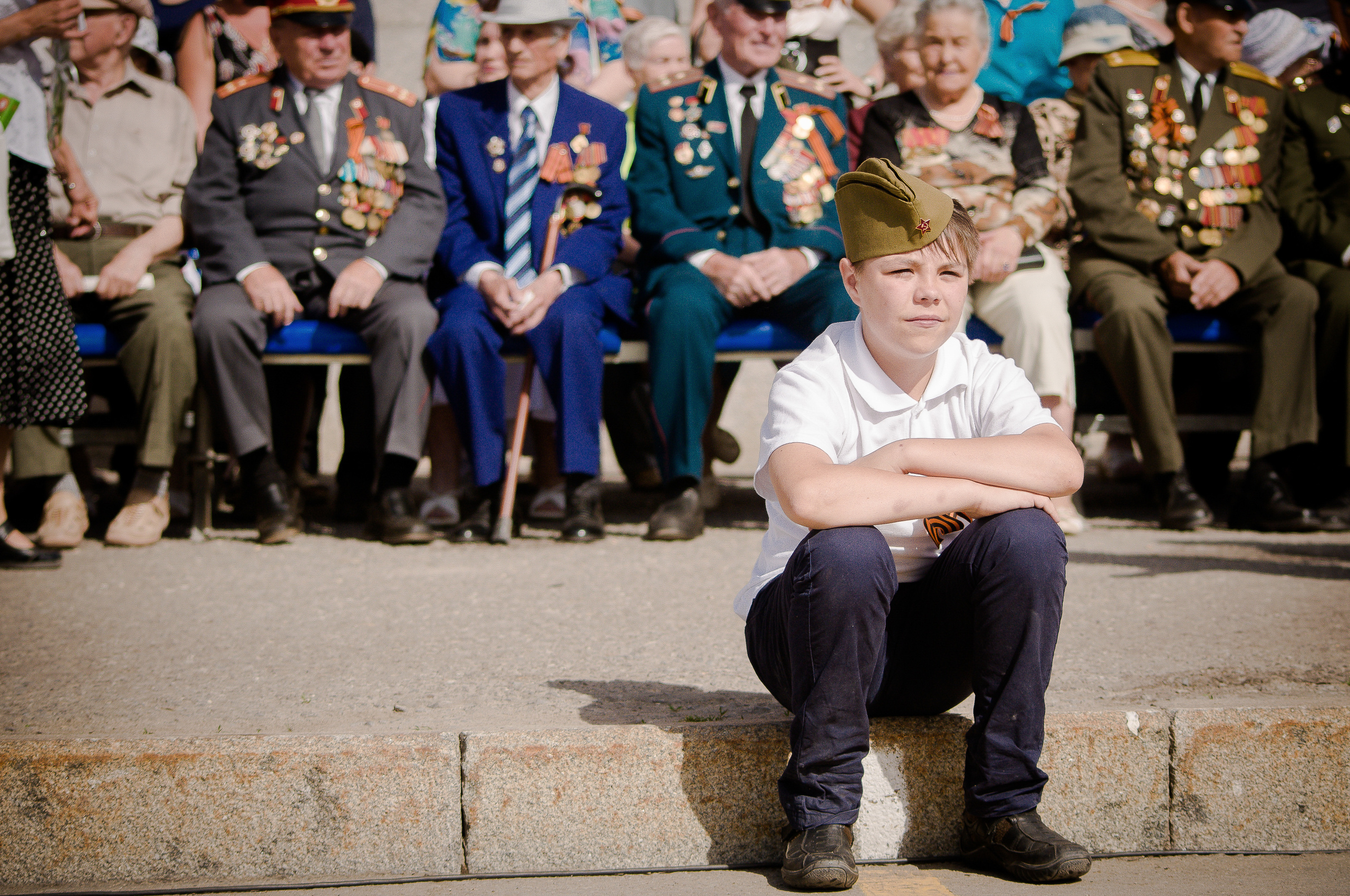 Ребёнок в пилотке сидит на земле на фоне ветеранов Великой Отечественной войны