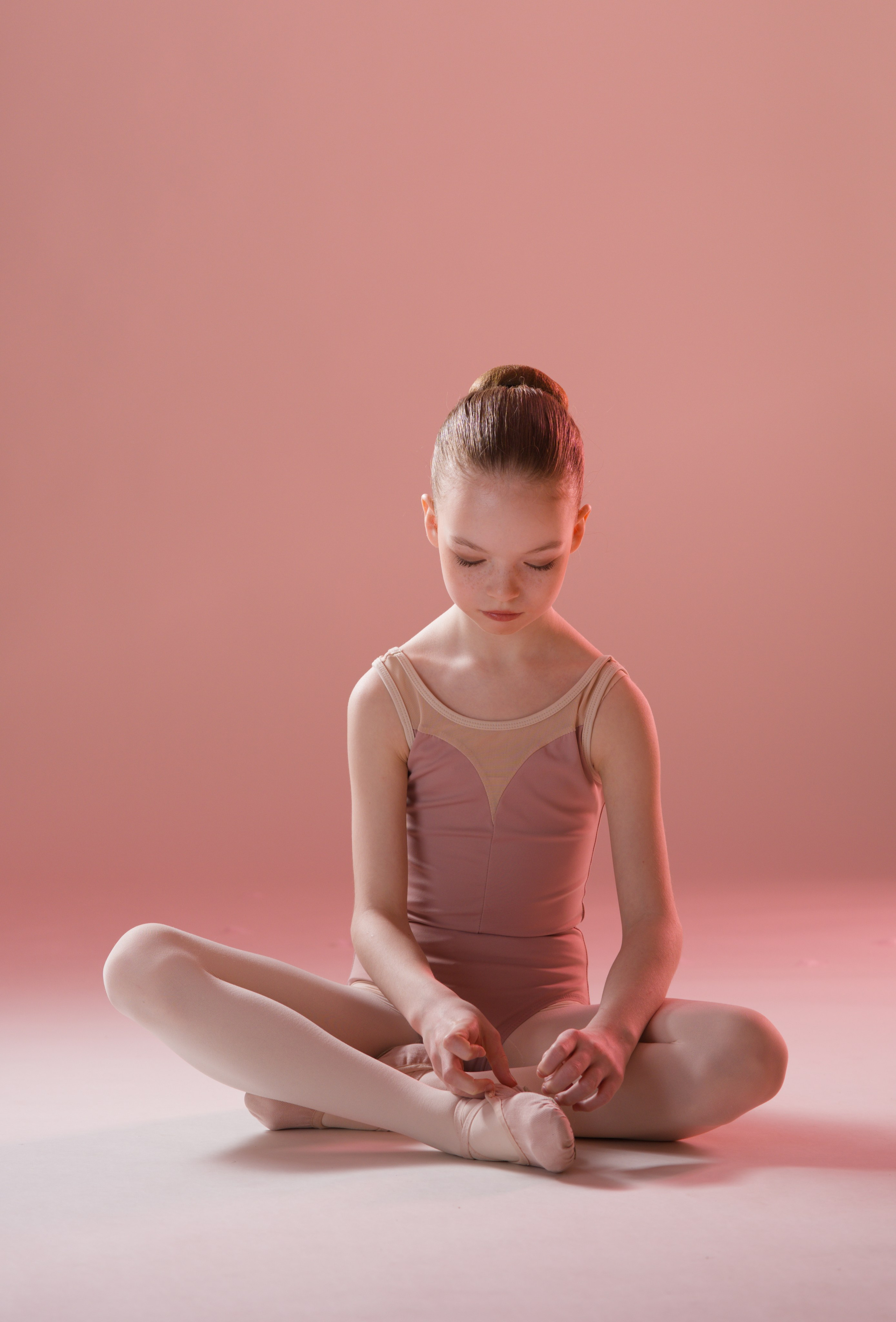Юная балерина сидит в лаконичной позе лицом к камере, мягкий розовый свет и минималистичный фон