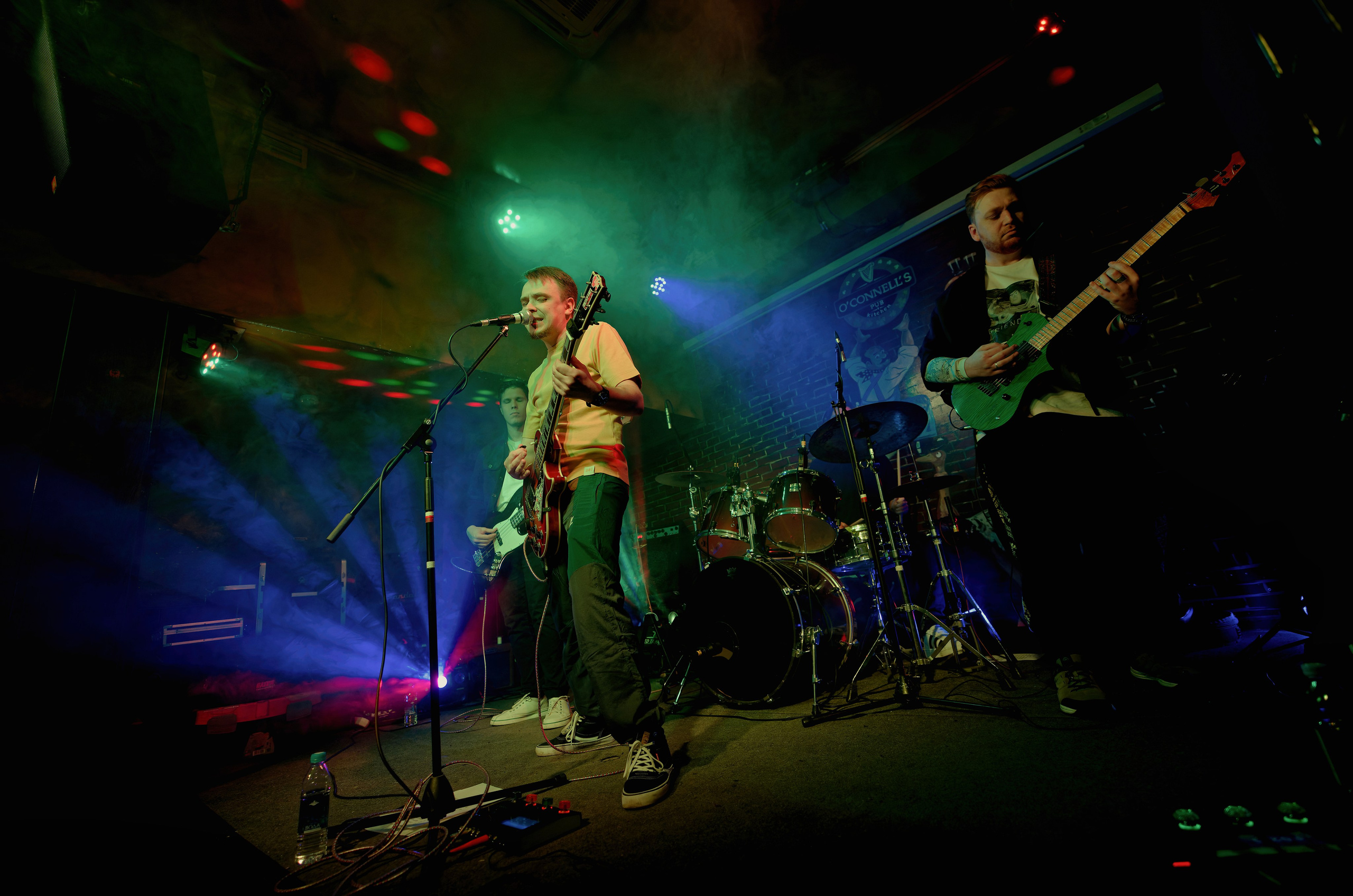 Рок-группа на сцене с цветным сценическим освещением и дымом - гитаристы и барабанщик, энергичное выступление, концертная фотография