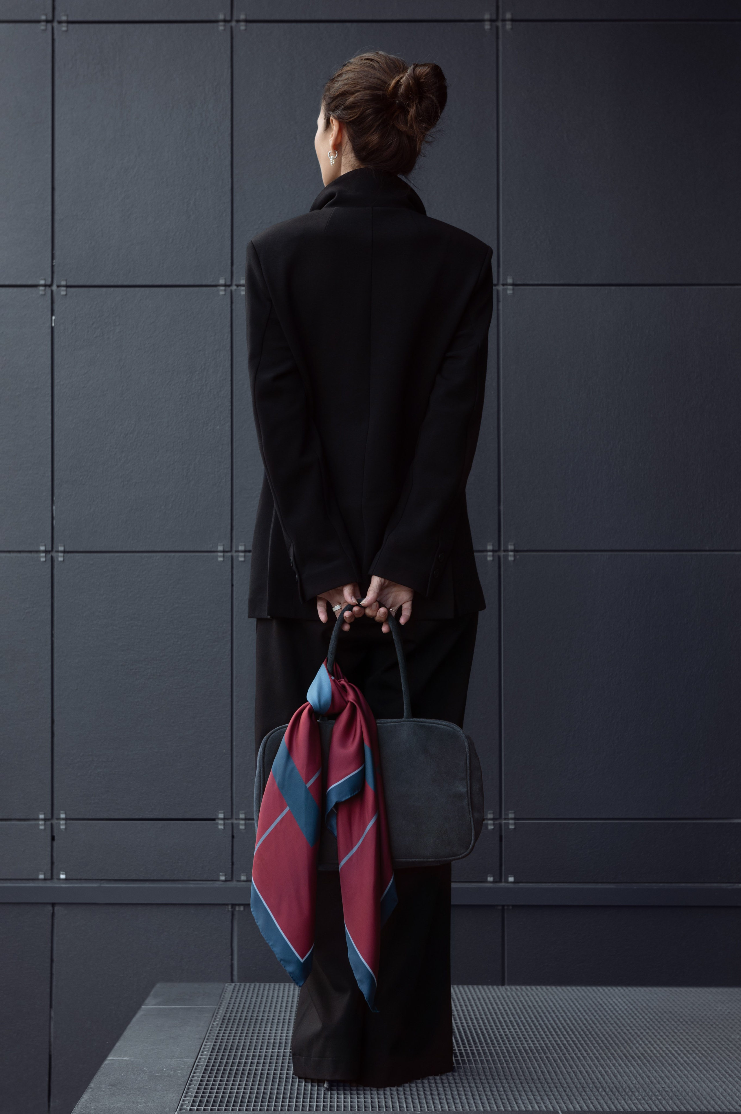 Девушка в черном брючном костюме стоит спиной к зрителю и держит сумку за спиной, на сумке повязан шелковый платок цвета бордо. Естественный свет падает сбоку на модель, красиво подчеркивая фактуру одежды. Съемка с естественным светом.