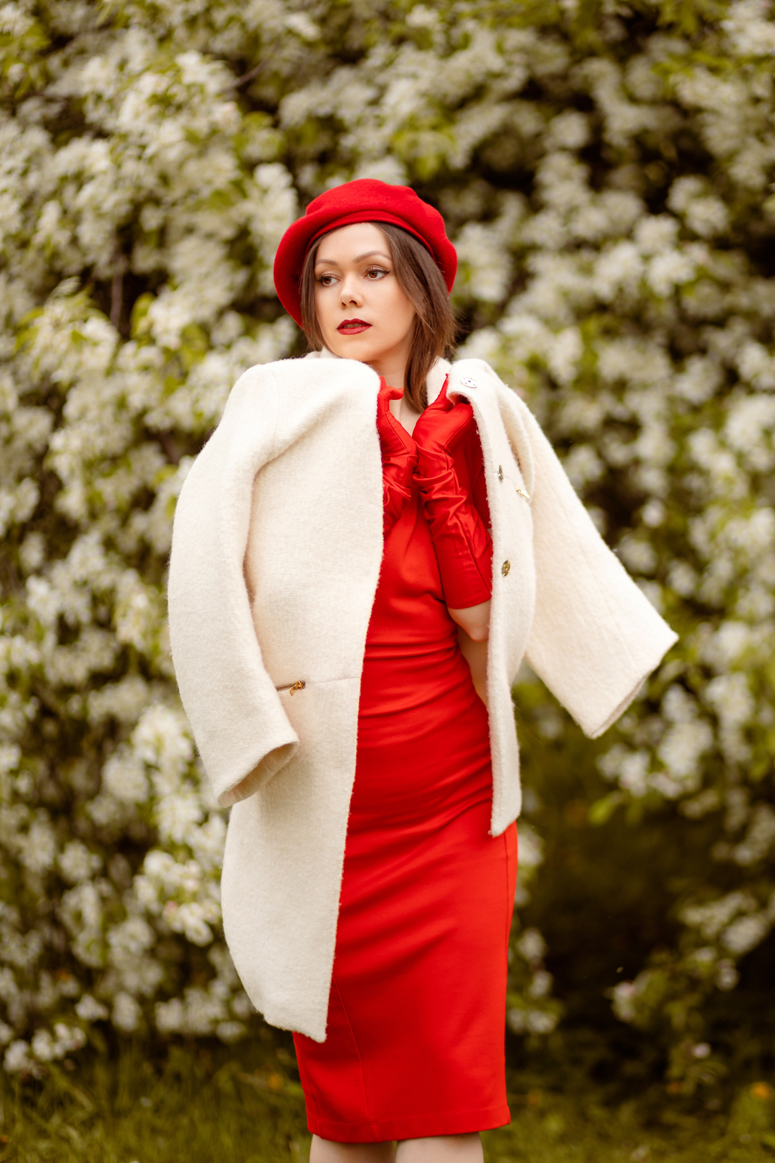 Девушка в белом пальто и красном платье позирует на фоне белого цветущего куста