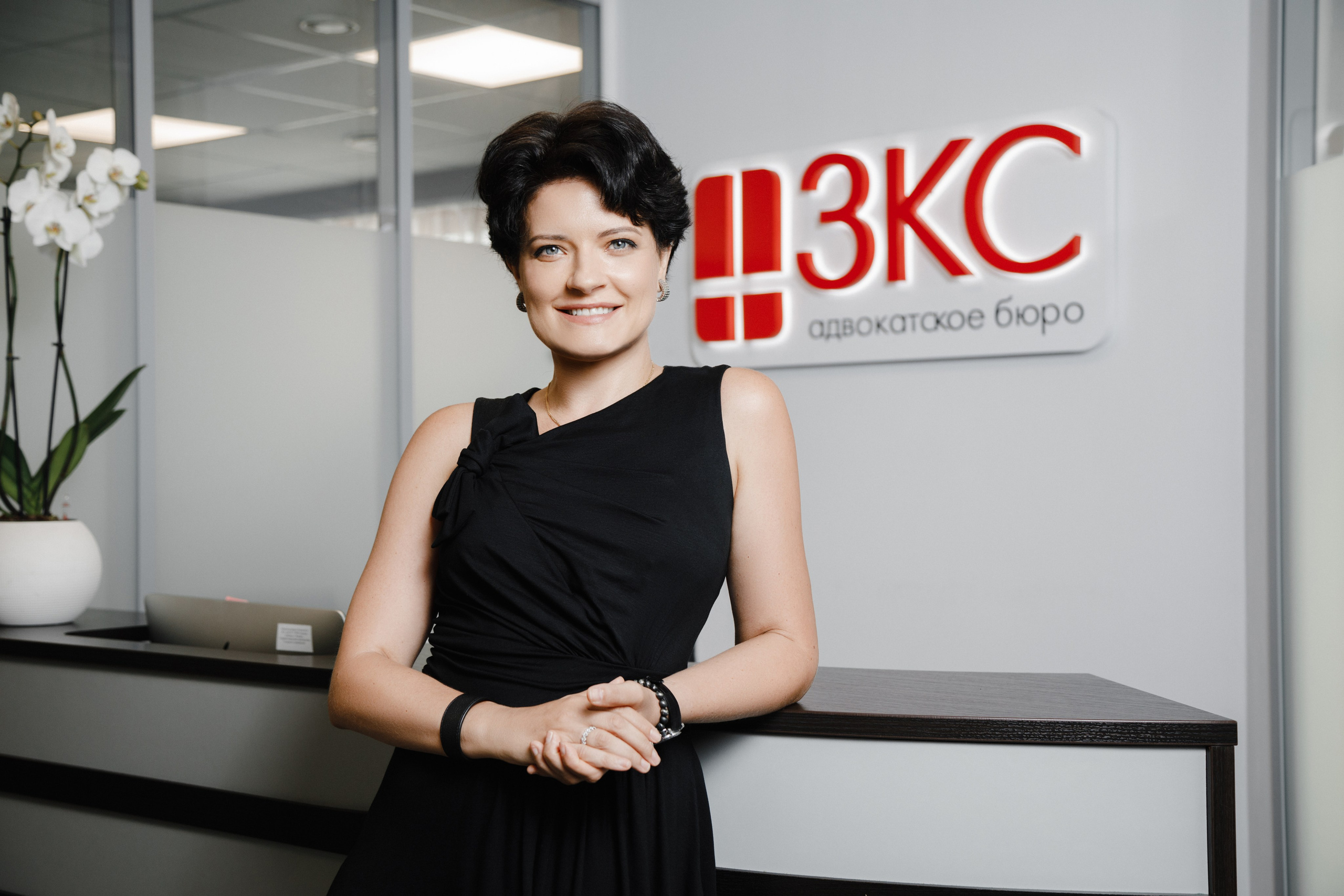 Девушка в деловой одежде позирует на фоне стойки ресепшена.