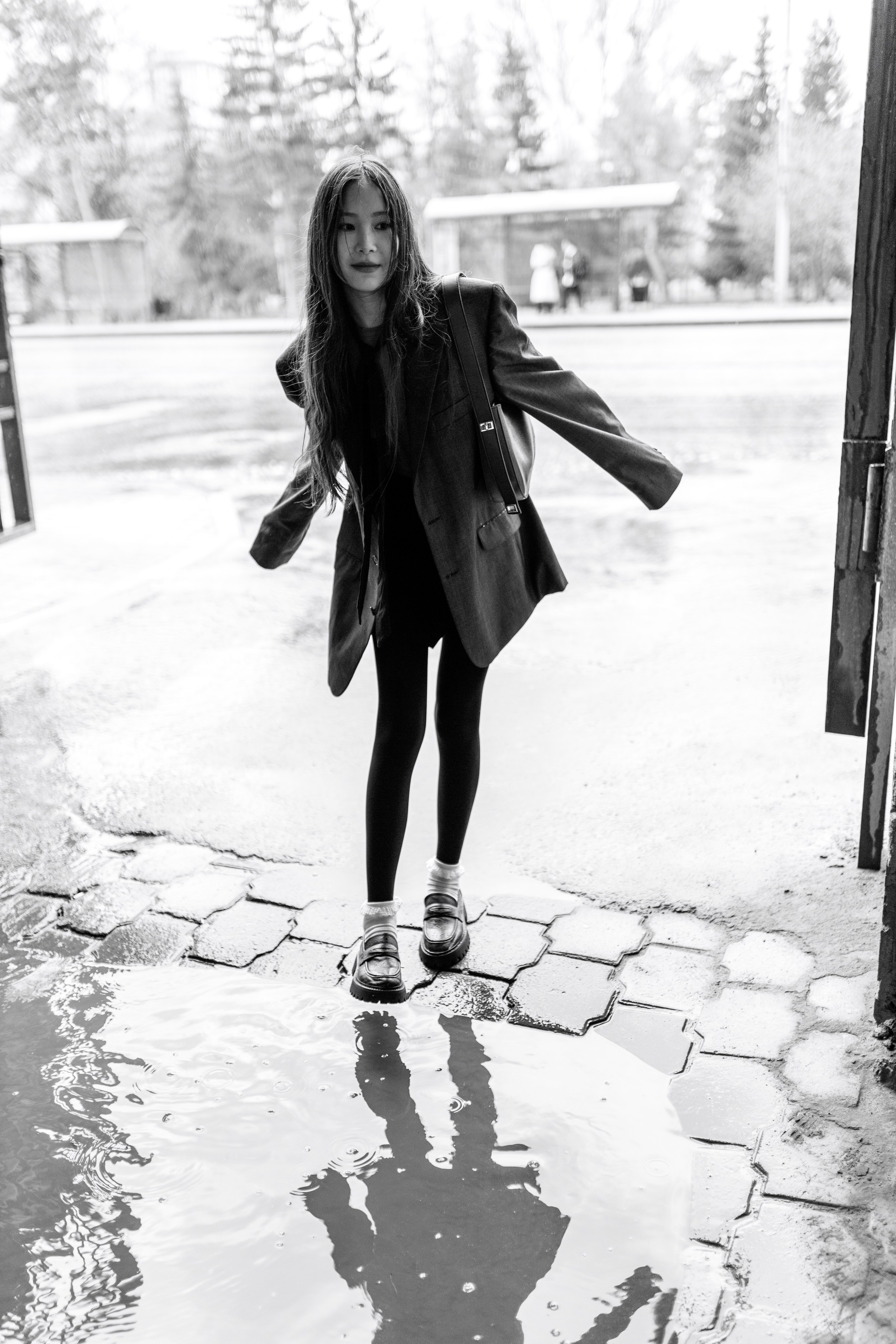 На краю большой лужи стоит молодая девушка в мешковатом пальто и леггинсах. В воде видно ее отражение, создающее игривую, беззаботную атмосферу.