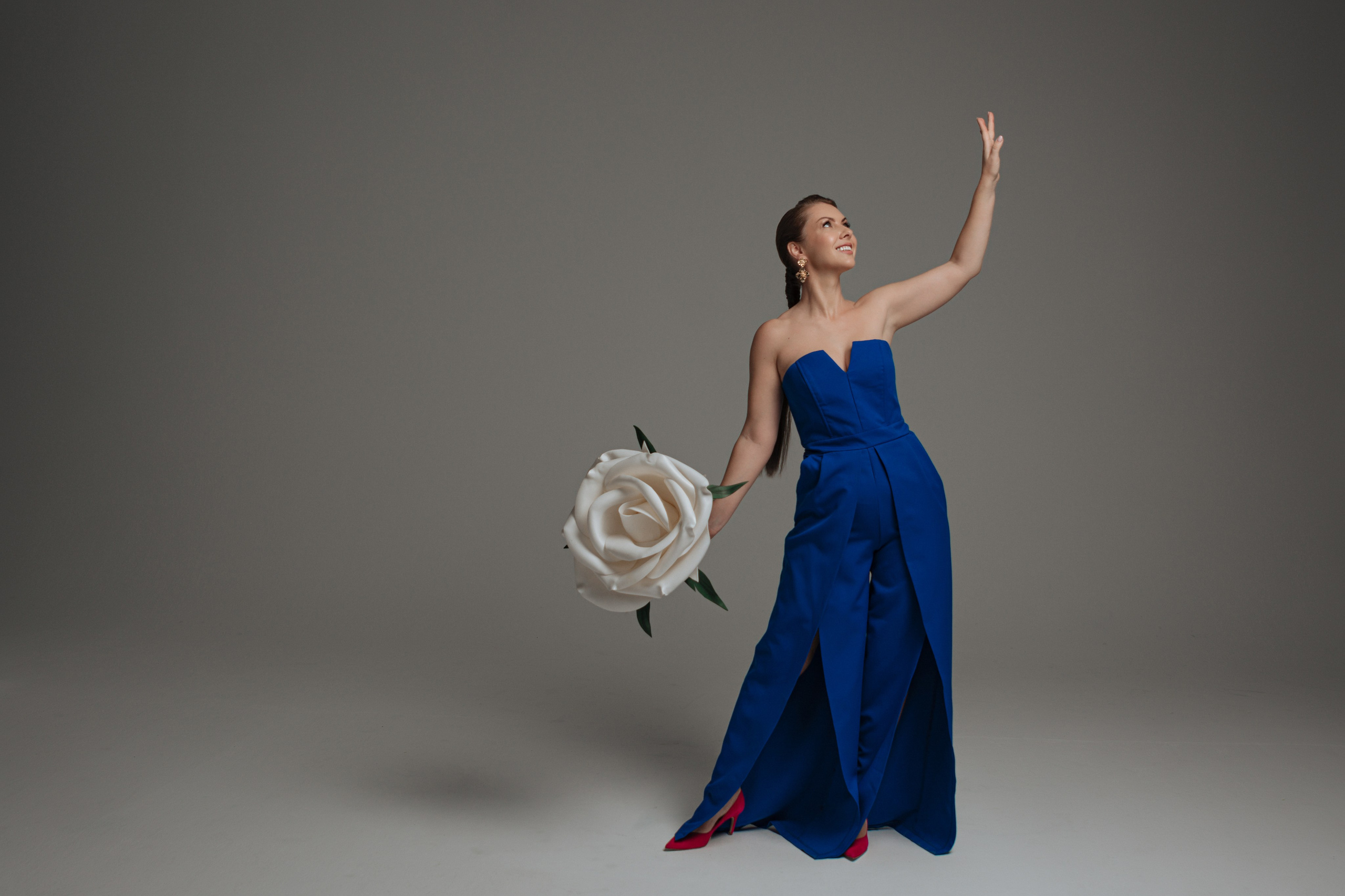 Ее струящееся платье цвета весеннего неба подчеркивает изящные изгибы фигуры, а широкий подол развевается при каждом шаге, будто лепестки распускающегося цветка. Контрастом роскошному синему наряду служит огромная белая роза, которую она бережно держит в руках. Пышные атласные лепестки цветка источают нежный, слегка пьянящий аромат, напоминающий о наступлении тёплых весенних деньков. 
