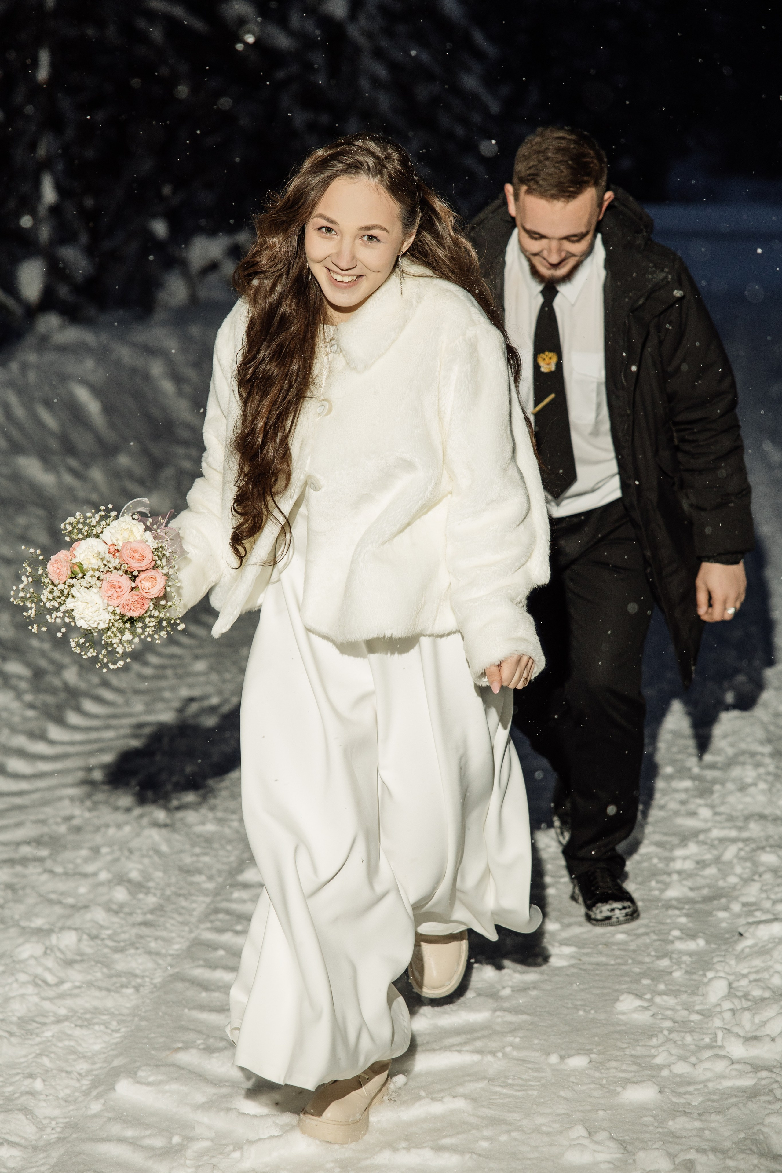 Живая зимняя свадебная фотосессия — невеста смеётся, жених идёт следом по снегу