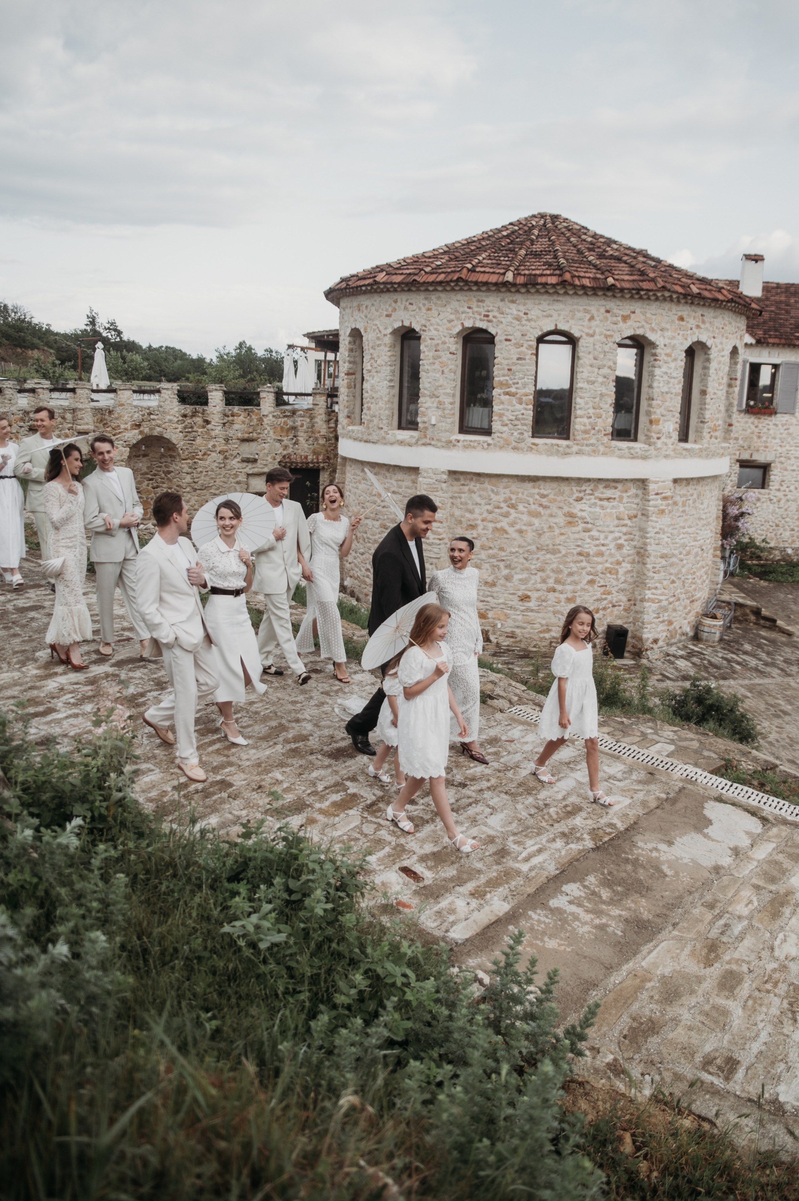 Свадебная процессия, состоящая из жениха, невесты и гостей, в светлых нарядах, идет по древним каменным дорожкам на фоне живописного здания