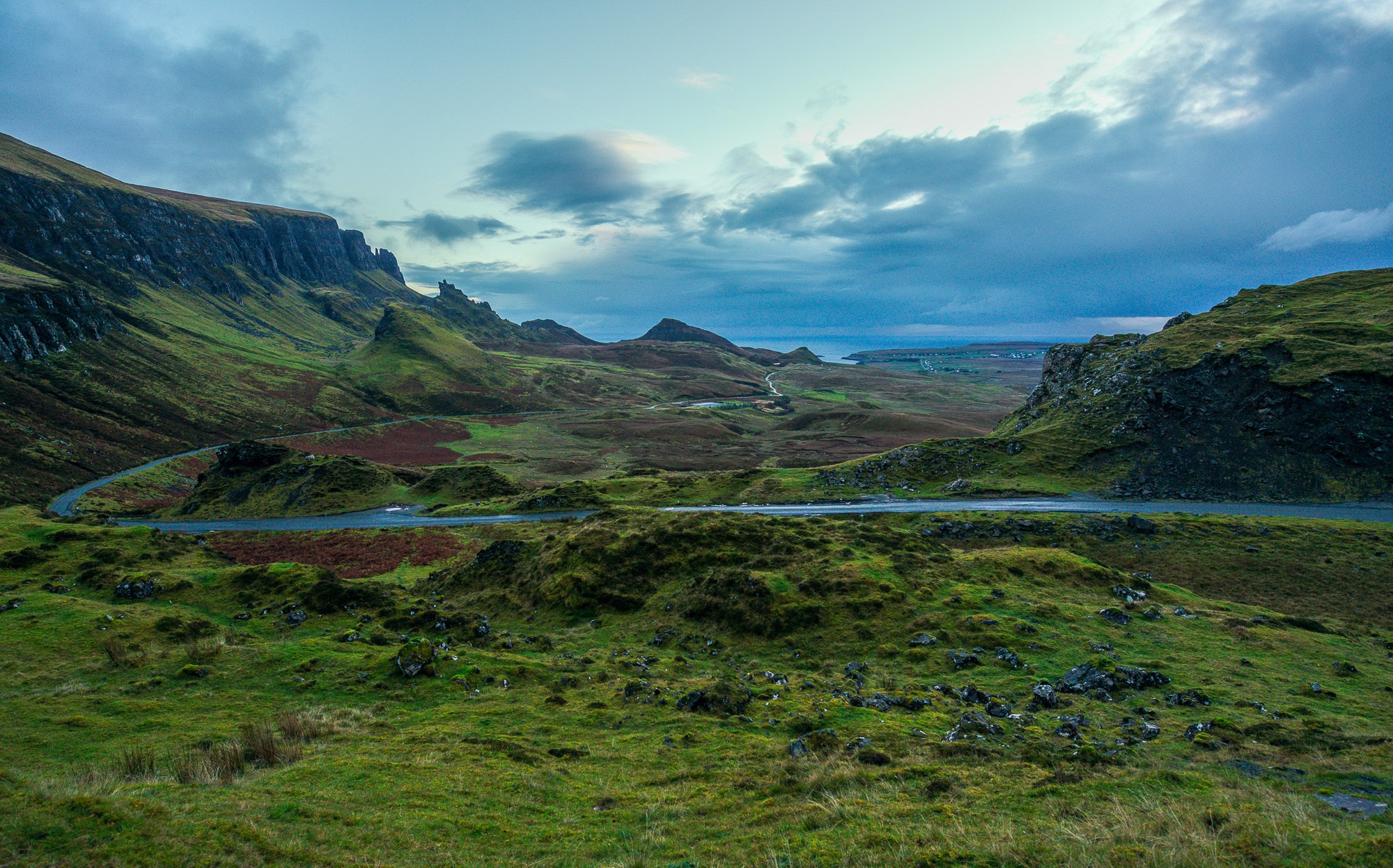 Панорамное фото перевала Квиранг на острове Скай в Шотландии: извилистая дорога на фоне драматичных зеленых гор, облачное небо и вересковые пустоши, атмосфера дикой природы