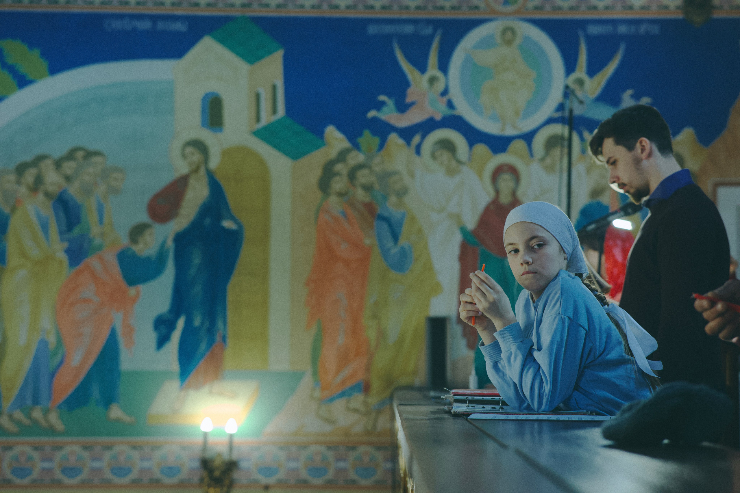 Девочка в голубом платке и мальчик стоят у аналоя на фоне большой фрески «Воскресение Христово». Девочка держит карандаш.