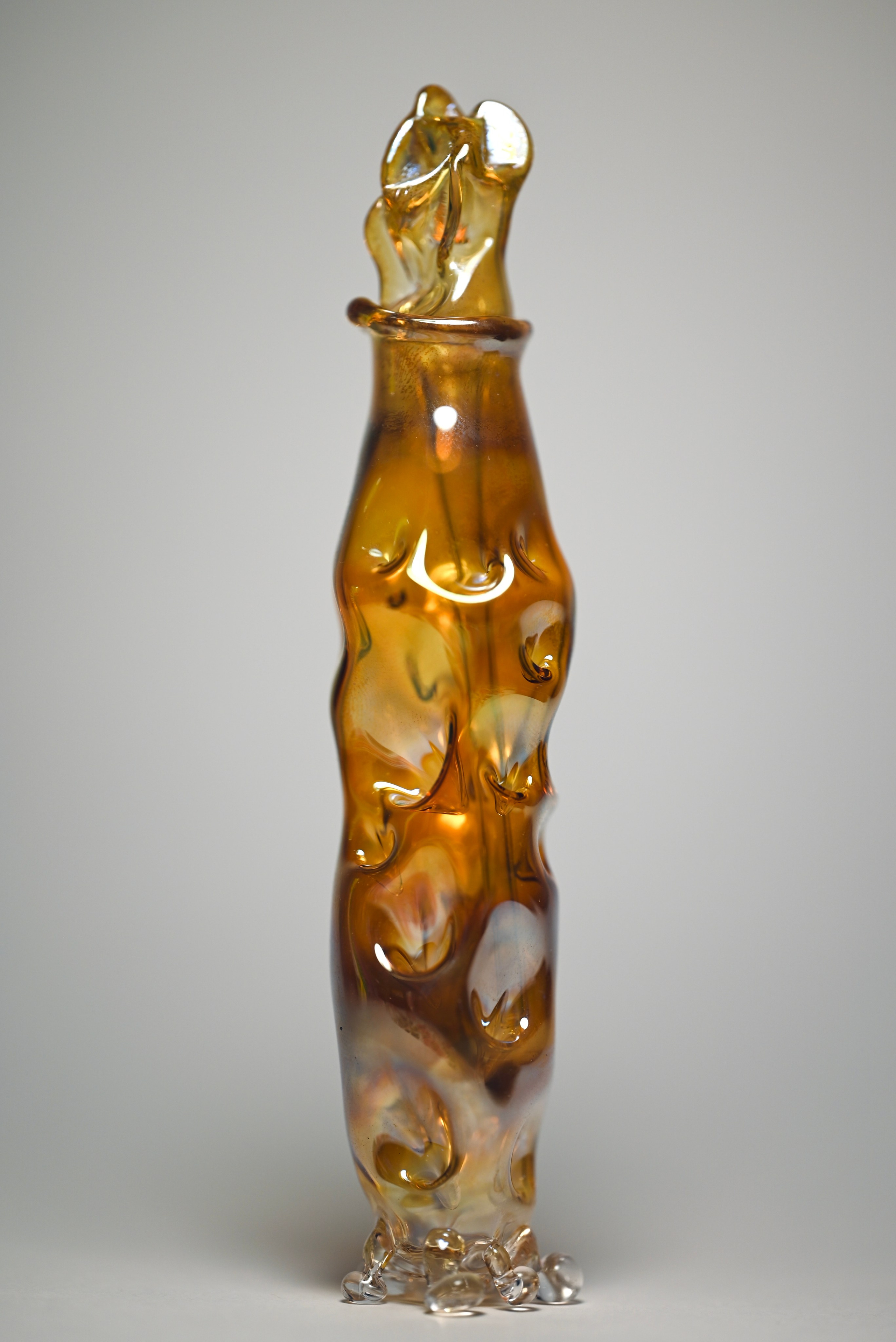 alt="Стеклянная скульптура в виде вытянутой бутылки с золотистым оттенком и округлыми выступами по всей поверхности."