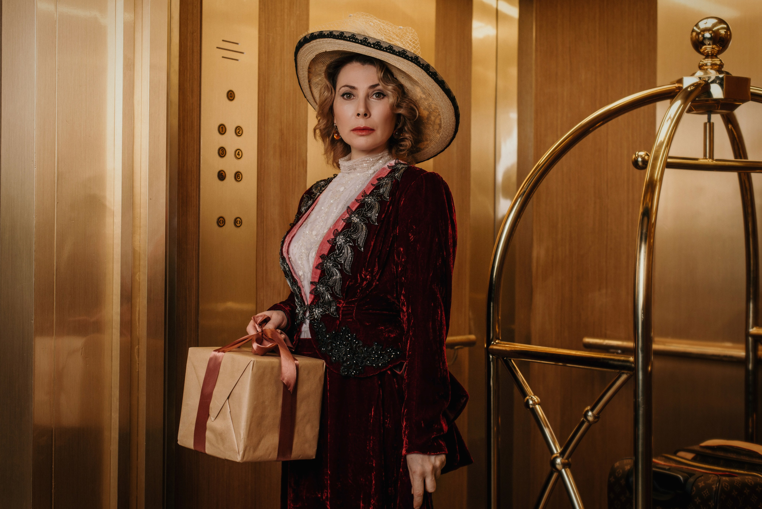 Фотосессия женщины в костюме и шляпе начала XX века в лифте отеля с подарком перевязанным лентой в руке