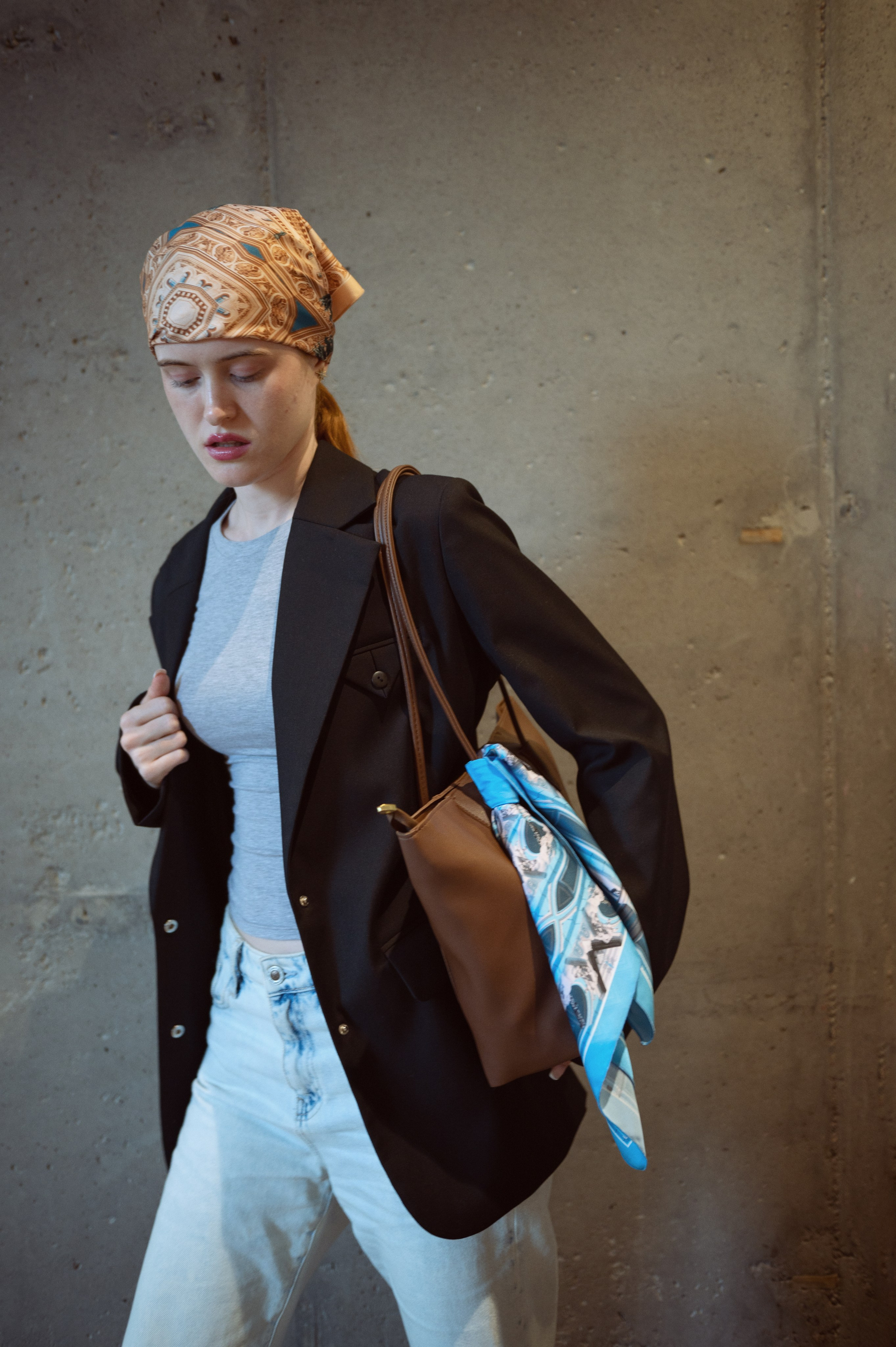 Красивая рыжеволосая девушка в ярко-желтом платке, черном пиджаке, серой майке и джинсах на фоне бетонной стены. На плече у нее коричневая сумка, украшенная голубым шелковым платком.