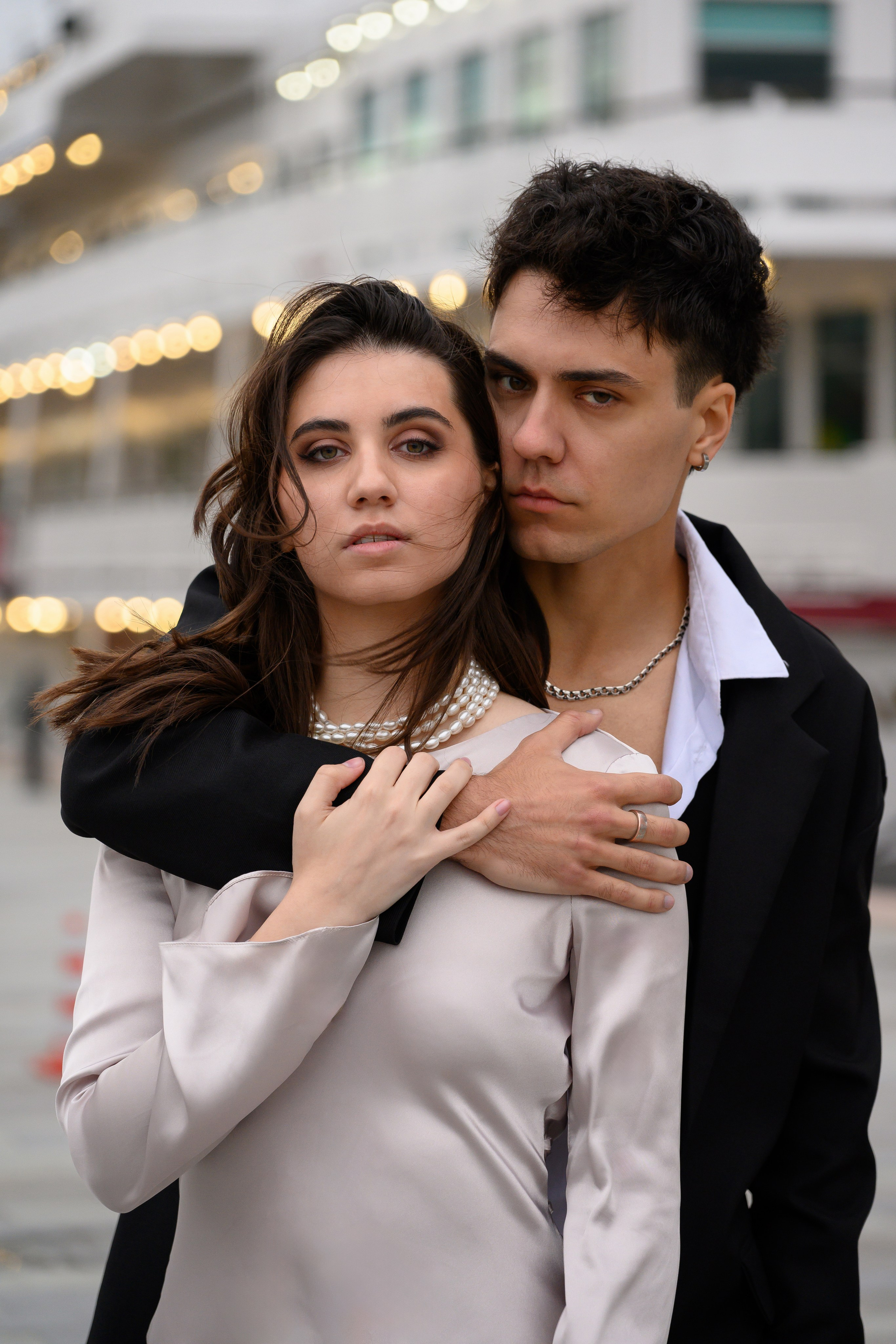 Молодой человек в белой рубашке и черном пиджаке обнимает за плечи стройную девушку в длинном струящемся платье.