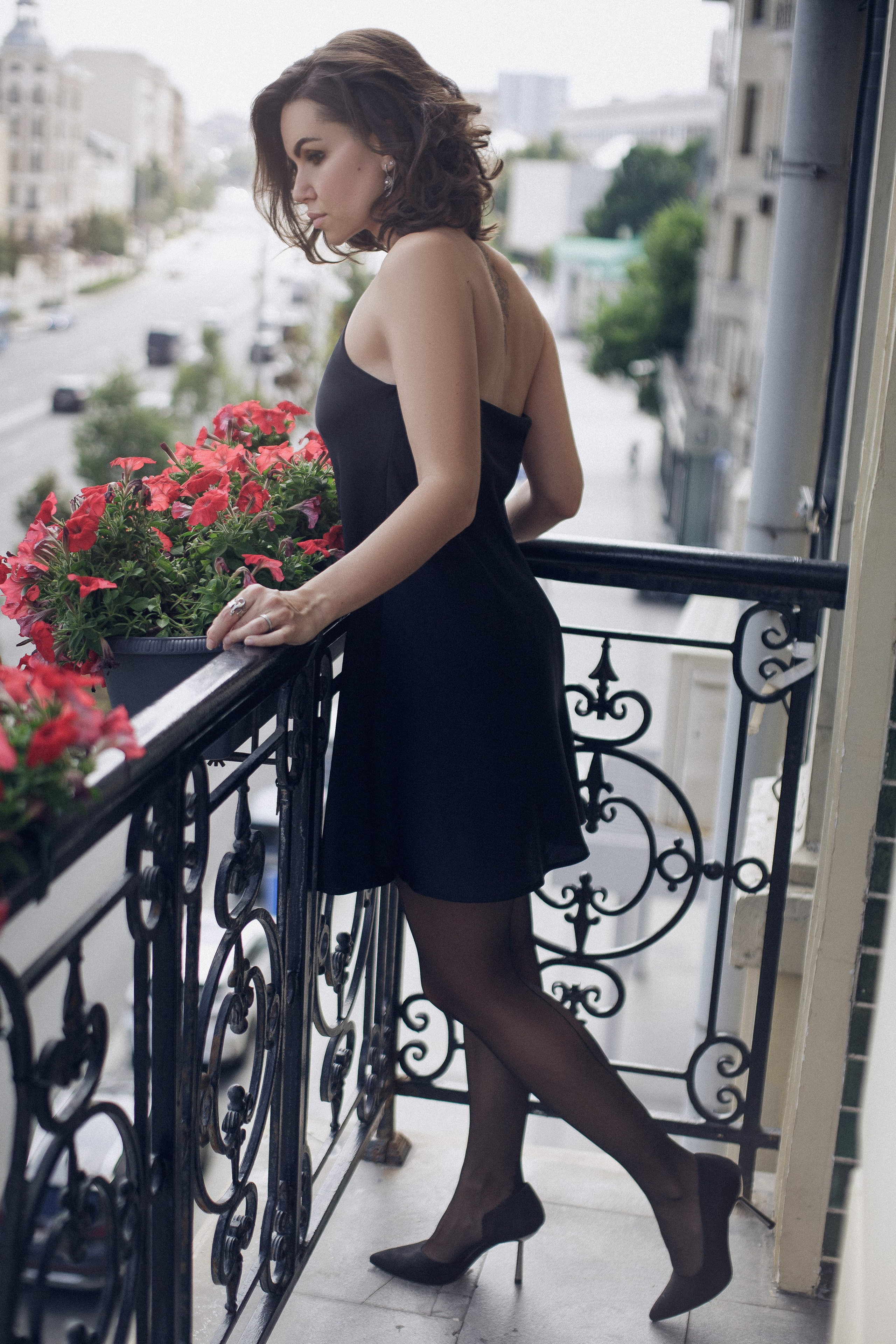 Фотография девушки в черном платье, которая стоит на балконе отеля с яркими цветами и видом на город.