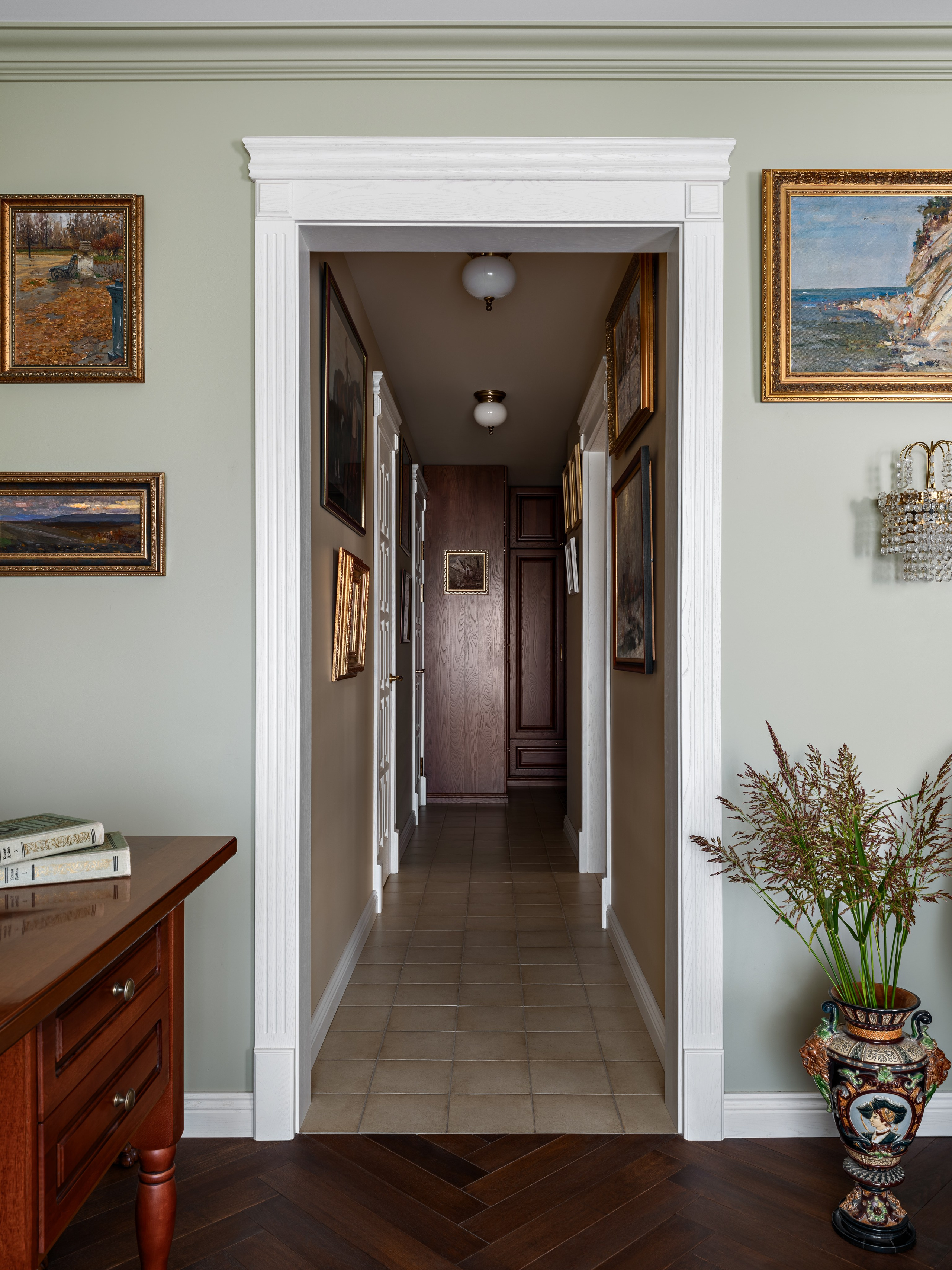 Элегантный интерьер коридора с мягким светом, картинами в золочёных рамах и классической белой дверной аркой; сочетание пастельных стен и тёплого дерева подчёркивает уют. Дизайн Юлии Капункиной и Альбины Терещенко, стиль Алины Нелень, фото Сергея Денисенко.