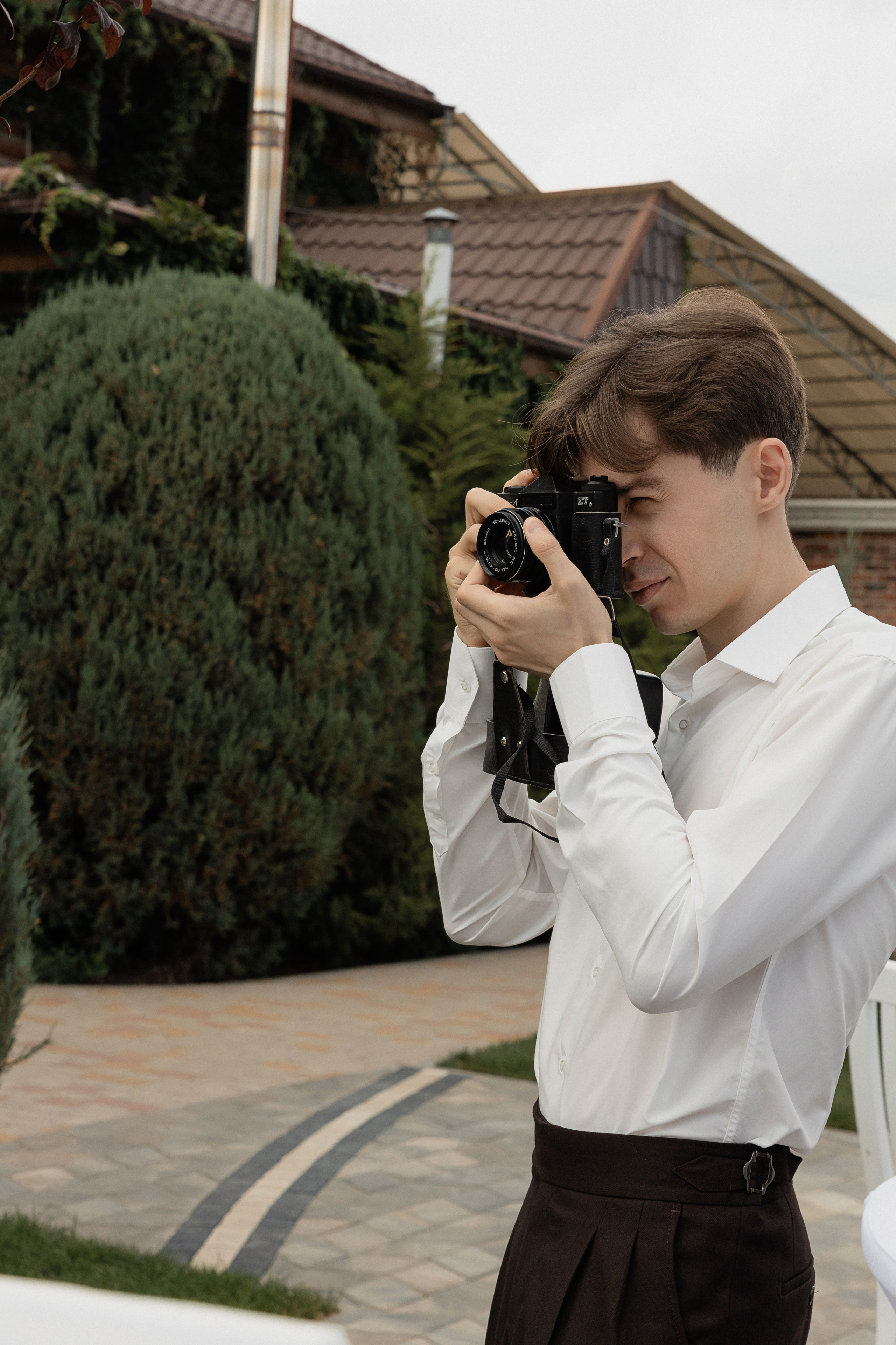 Жених на свадьбе в белой рубашке фотографирует гостей на плёночный фотоаппарат во дворе банкетного зала Шодо