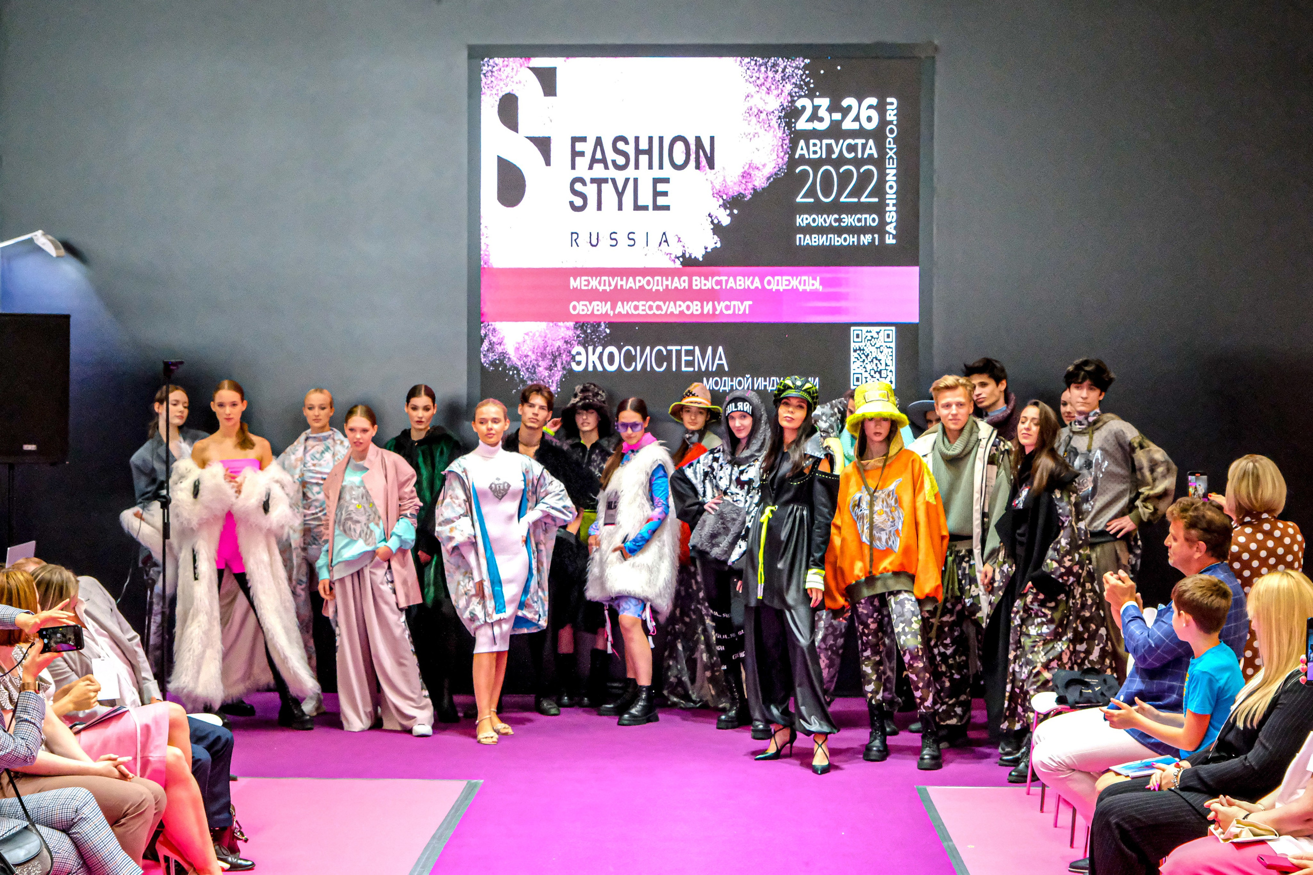  "Модели в ярких нарядах проходят по подиуму во время показа мод, освещенные яркими софитами и окруженные восторженными зрителями."