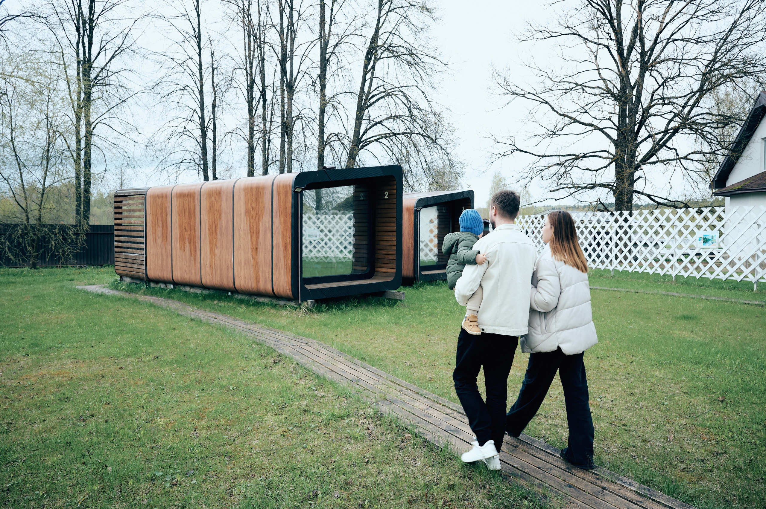 Семья идёт по дорожке к модульному деревянному домику на фоне весеннего пейзажа