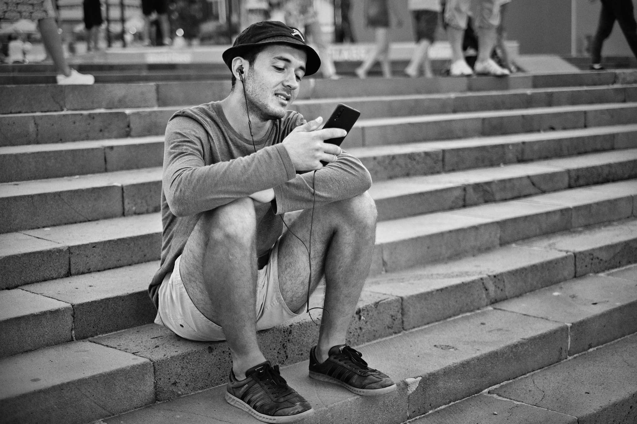 Embankment near the seaport of Sochi. A guy sits on the steps of the embankment contemplating his smartphone with a joyful expression on his face. Behind him are the legs of people passing by. (Набережная возле морского порта Сочи. Парень сидит на ступенях набережной созерцая свой смартфон с радостным выражением на лице. Позади него видны ноги людей проходящих мимо.)