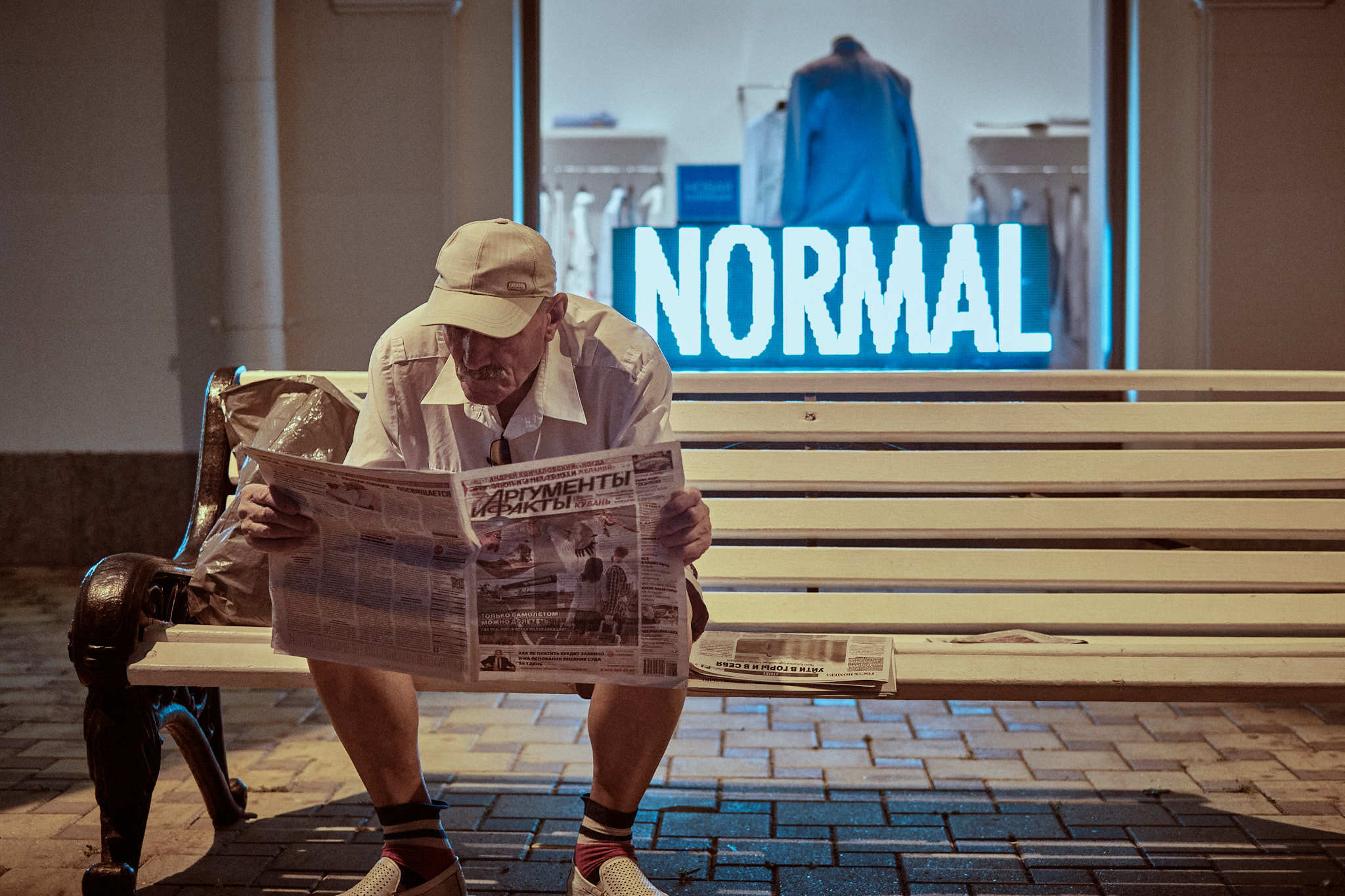 Sochi sea port. An elderly man is reading the newspaper "Arguments and Facts" sitting on a bench. In the background behind it, a shop window is visible, in which the inscription "NORMAL" glows. (Сочинский морской порт. Пожилой мужчина читает газету "Аргументы и Факты" сидя на скамейке. На фоне за ним видна витрина магазина, в которой светится надпись "NORMAL".)