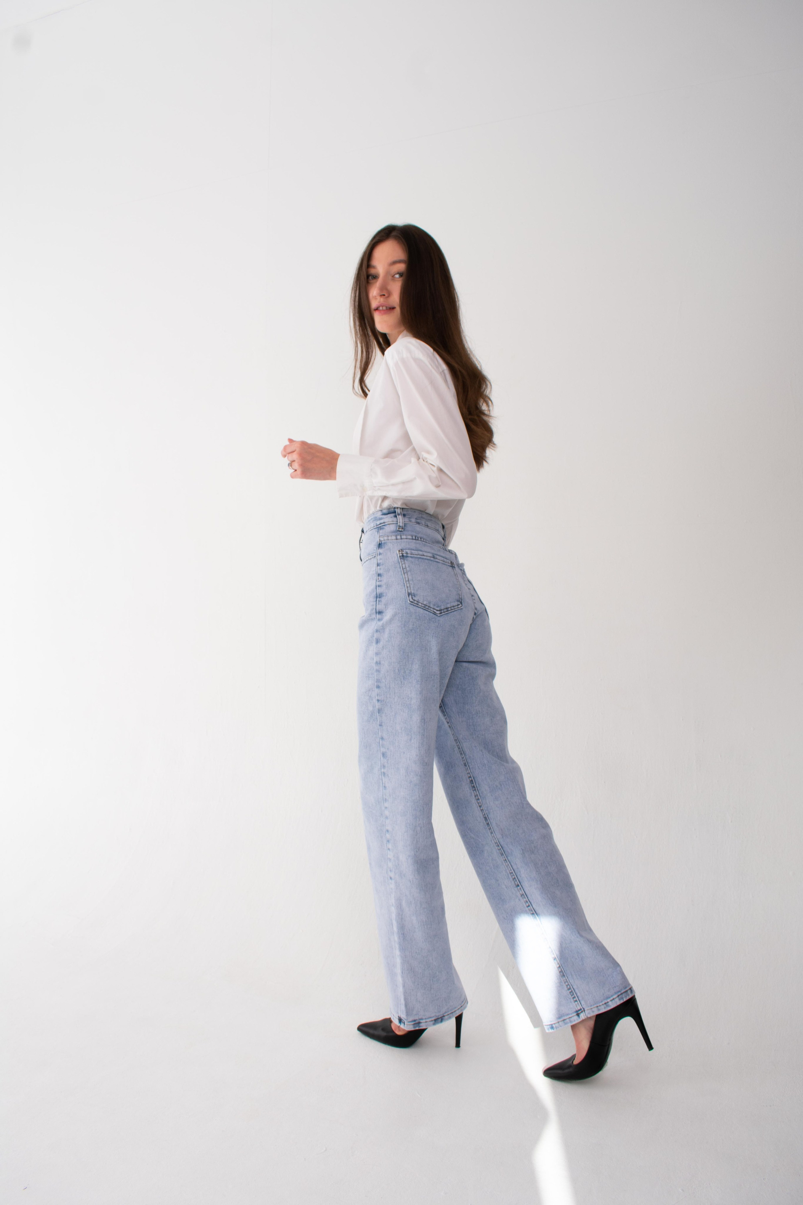 Модель фотографируется в студии в белой рубашке, джинсах и каблуках