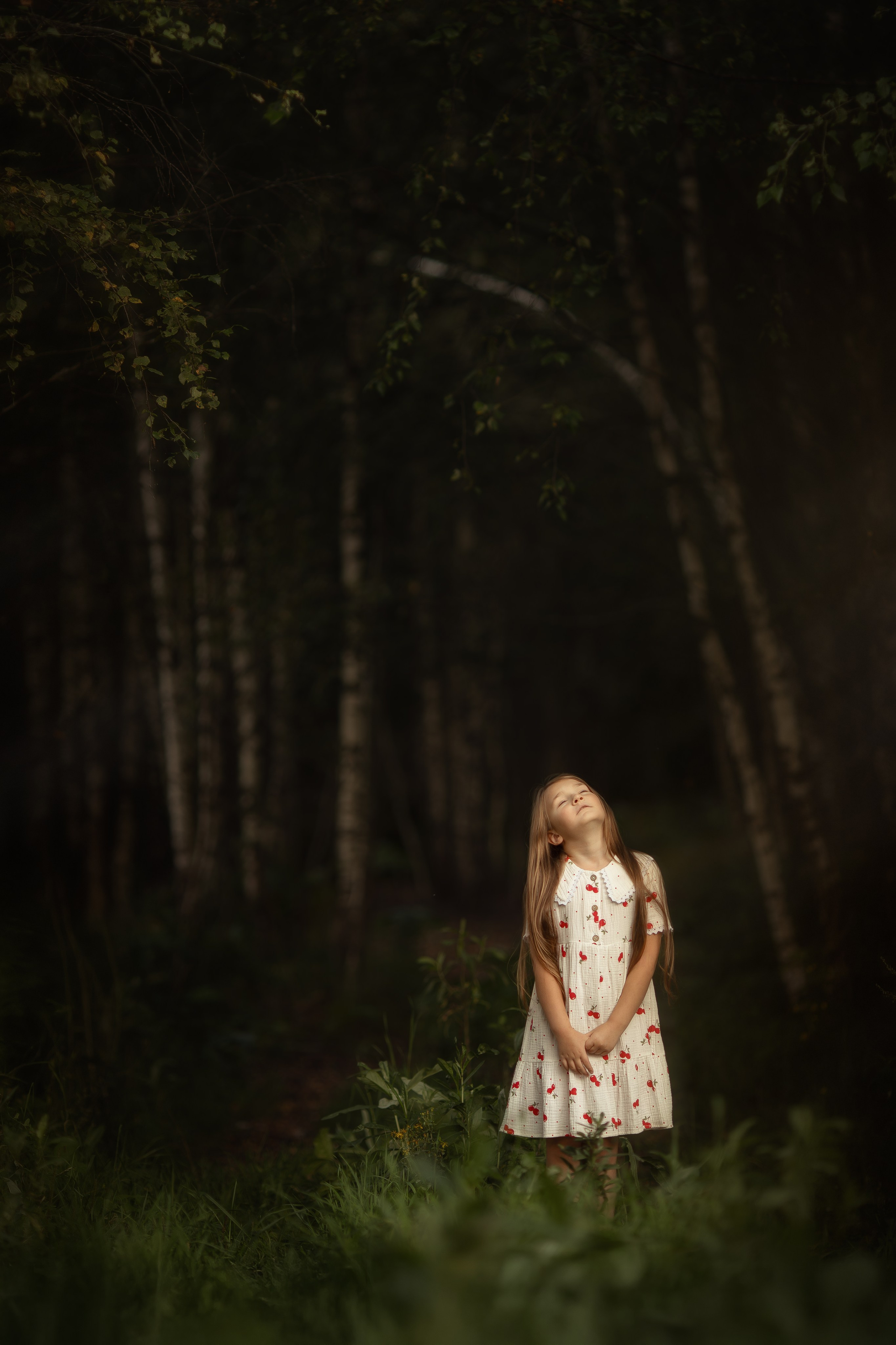 Девочка в белом платье с красным орнаментом стоит в тёмном лесу, на неё падает тёплый солнечный луч, вокруг густые деревья и тишина.