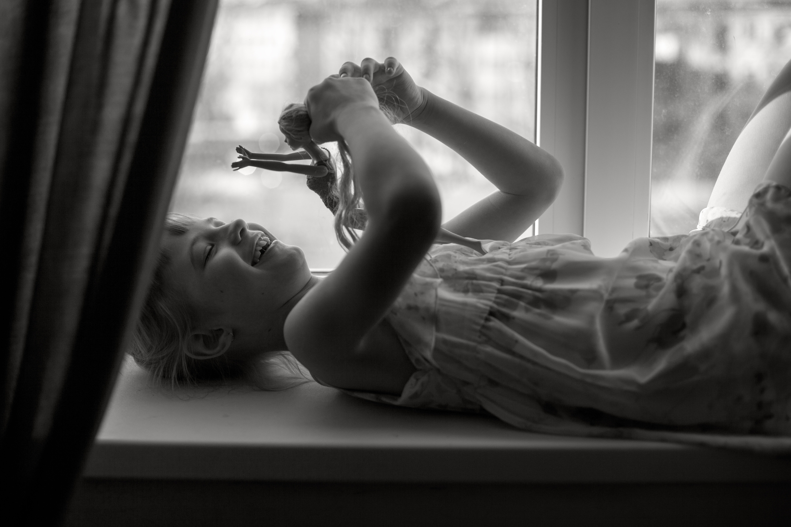 Девочка играет с куклой на окне, весёлая. Лучи солнца светят на её тело.