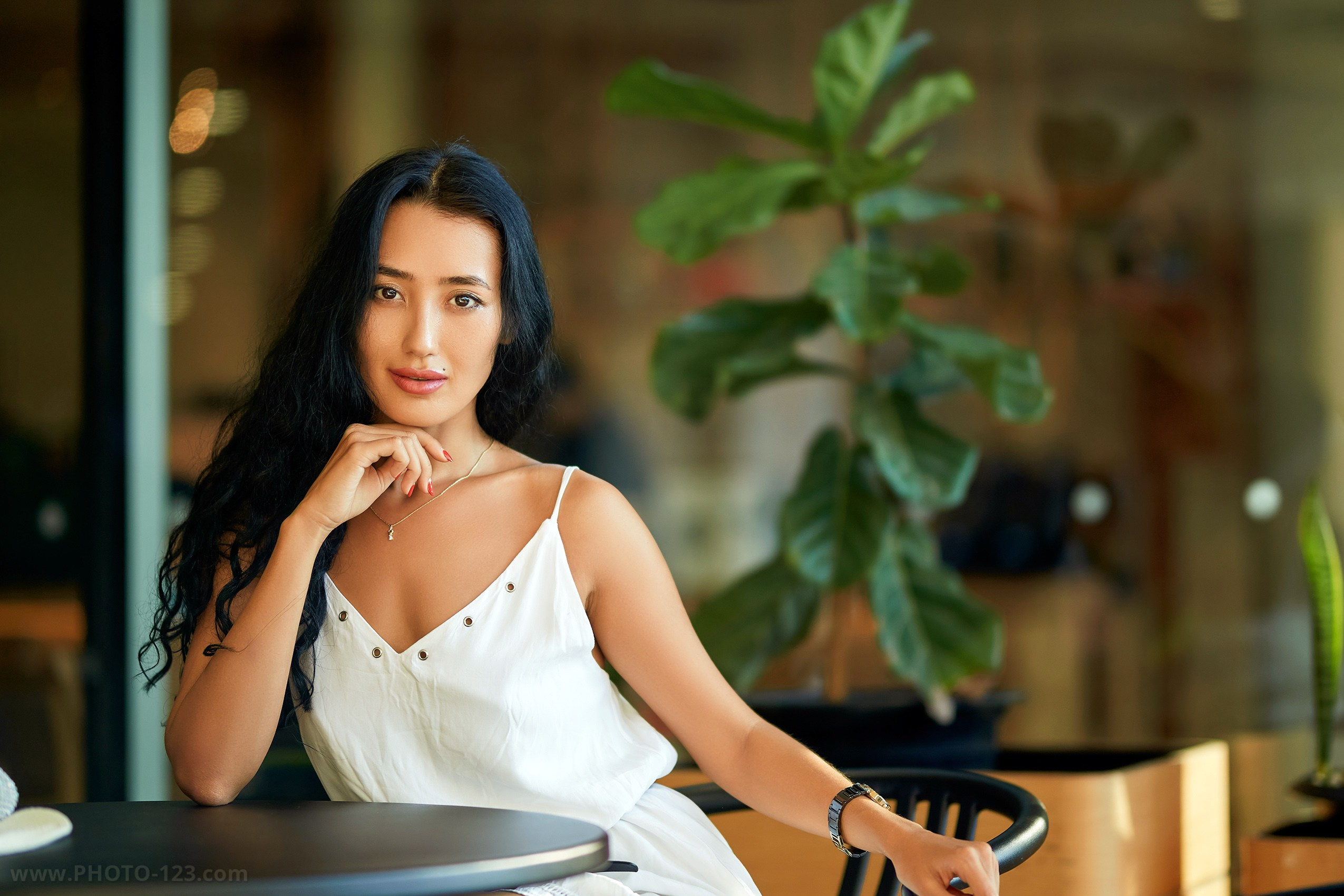 Portrait of a woman in a white dress seated at a café table, natural light and indoor plants in the background, photographed by a professional fashion photographer