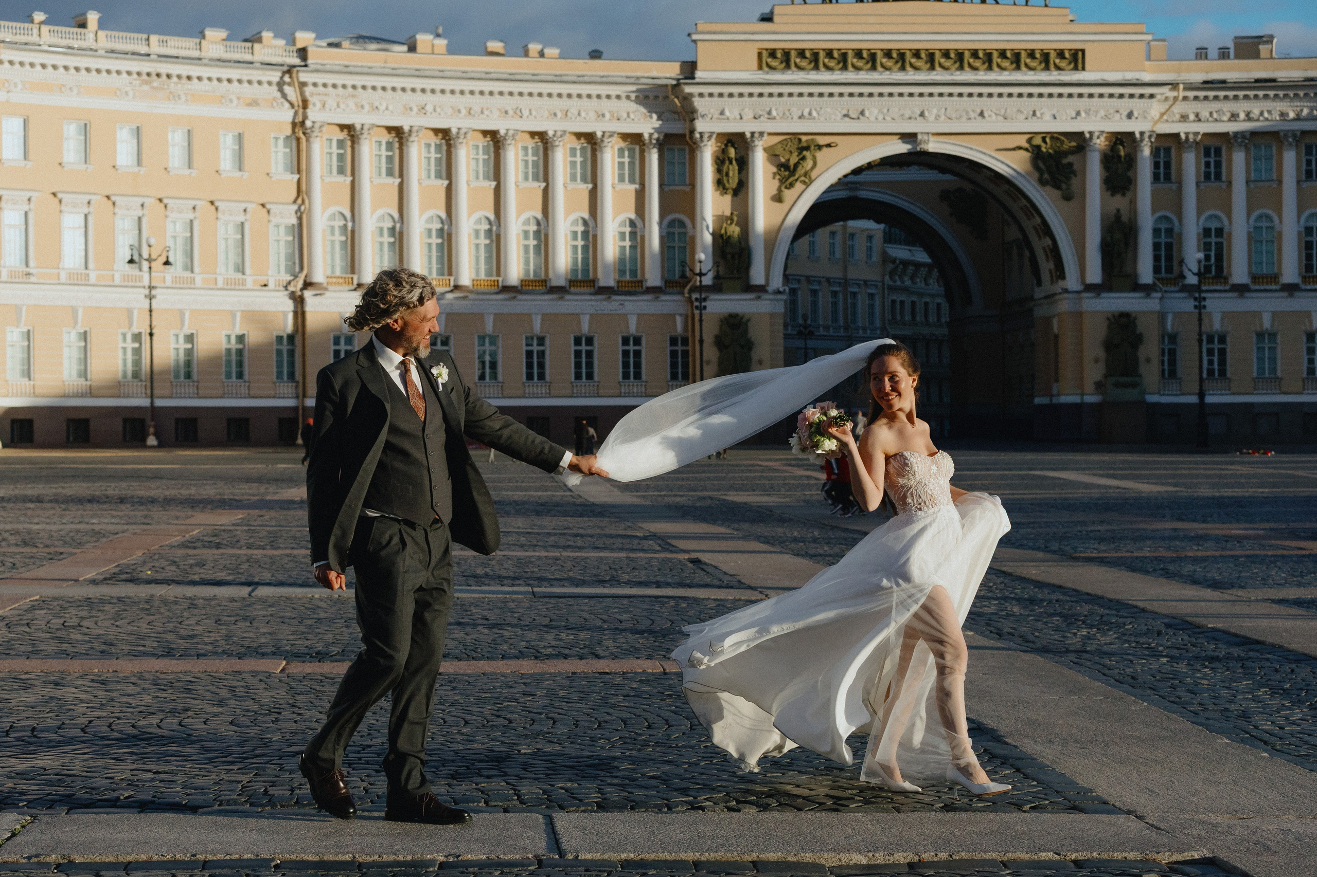 Свадебная фотосессия на Дворцовой площади Санкт-Петербурга: жених и невеста идут, ветер развевает фату.