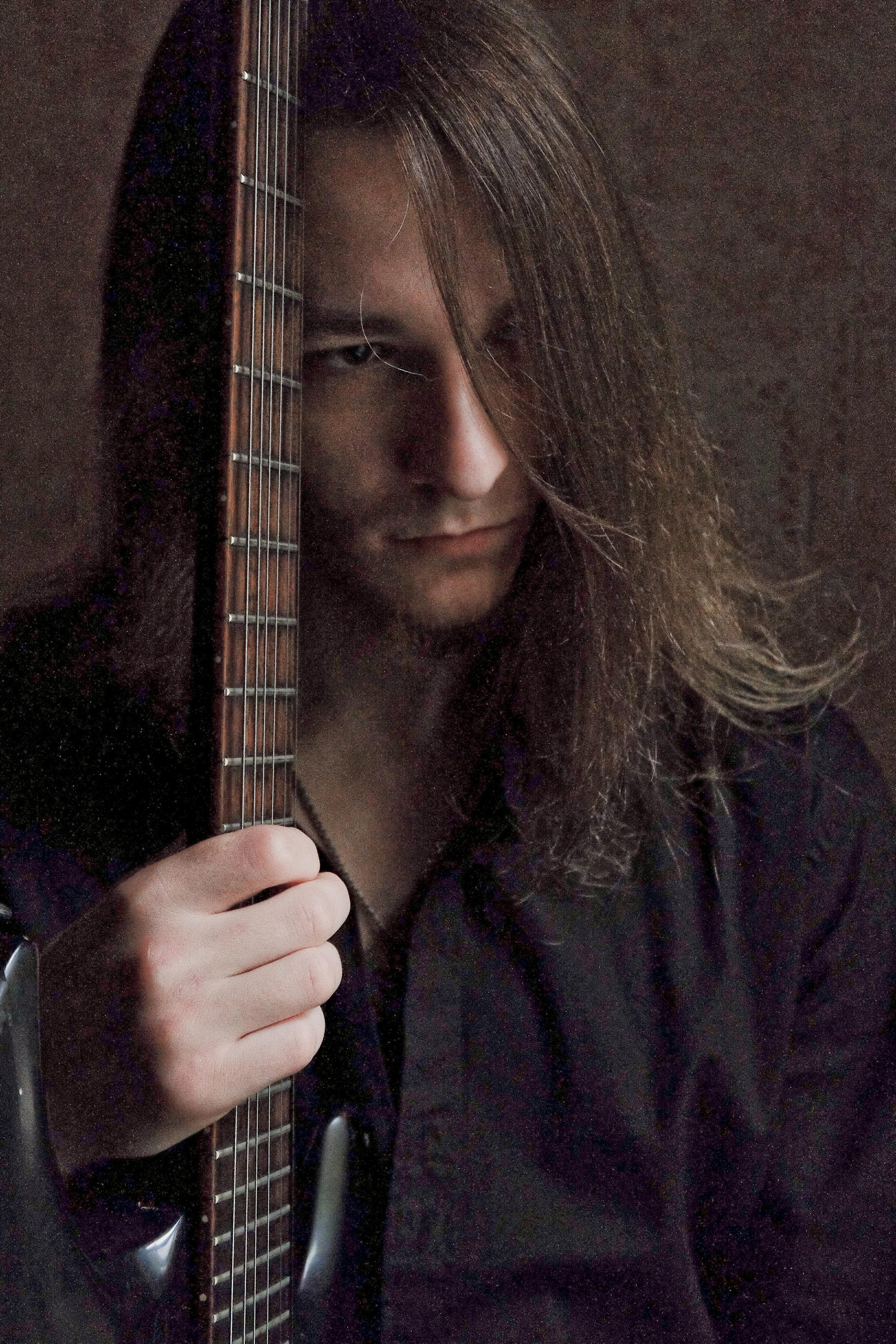 молодой человек музыкант с длинными волосами прижимает электрогитару грифом к голове