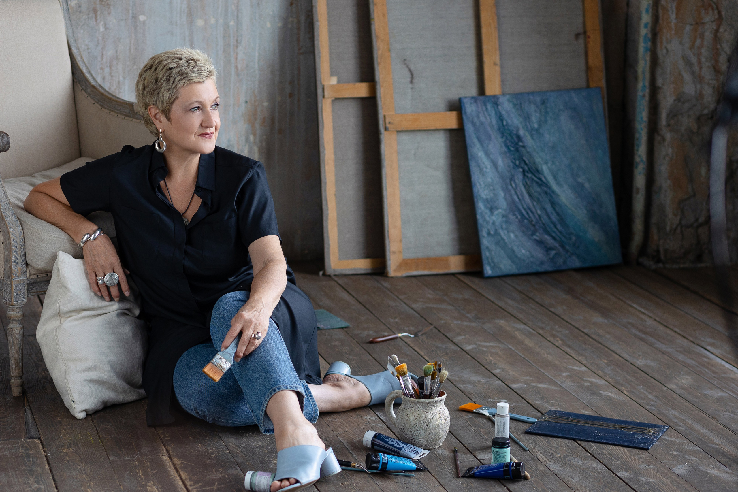 Художница сидит на полу облокотившись на кресло, слева от нее у стены стоят холсты картин.