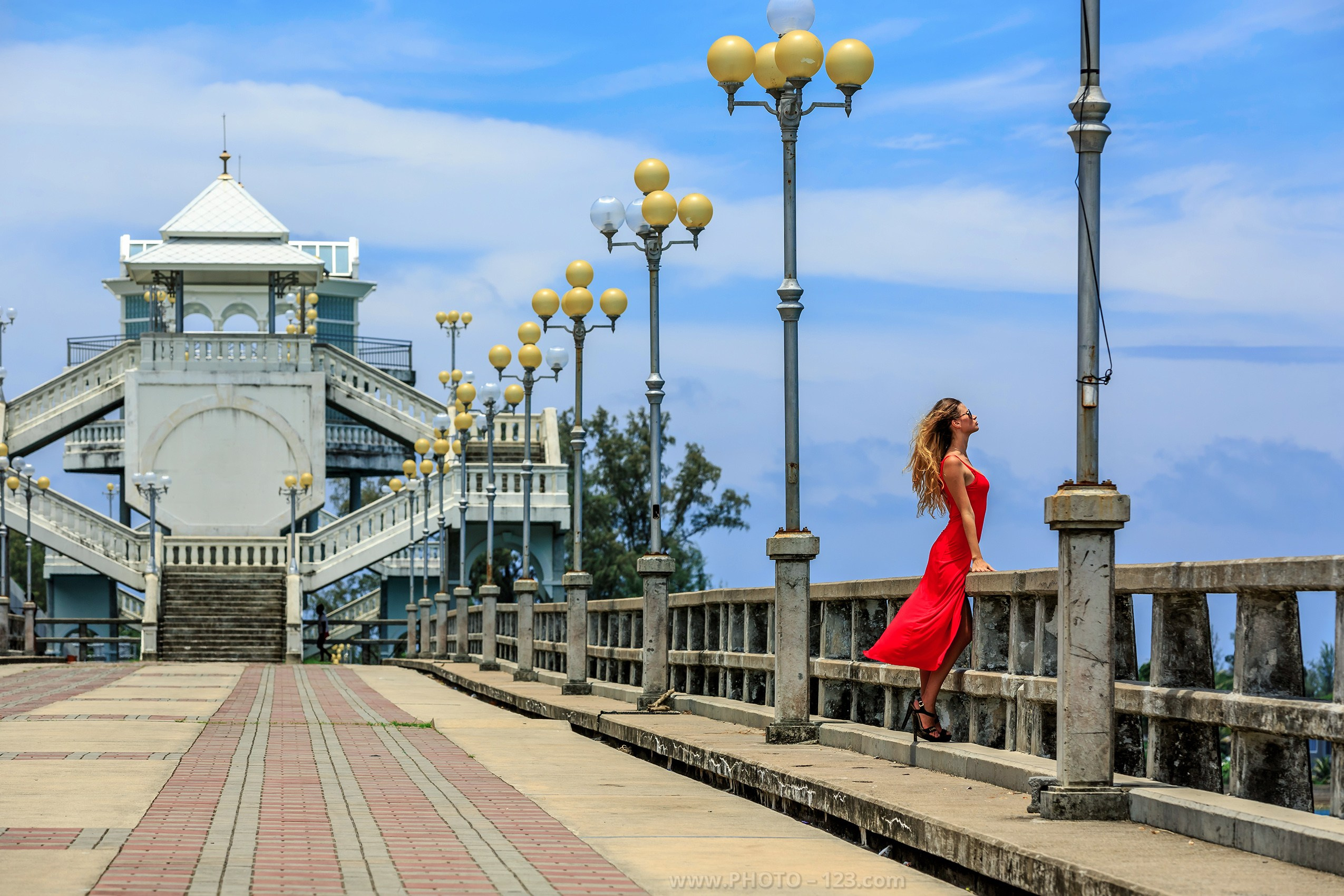 Woman in red dress standing on city promenade bridge, architectural symmetry with street lamps, blue sky, elegant posture, minimal urban scene, sense of calm and freedom, travel and lifestyle concept